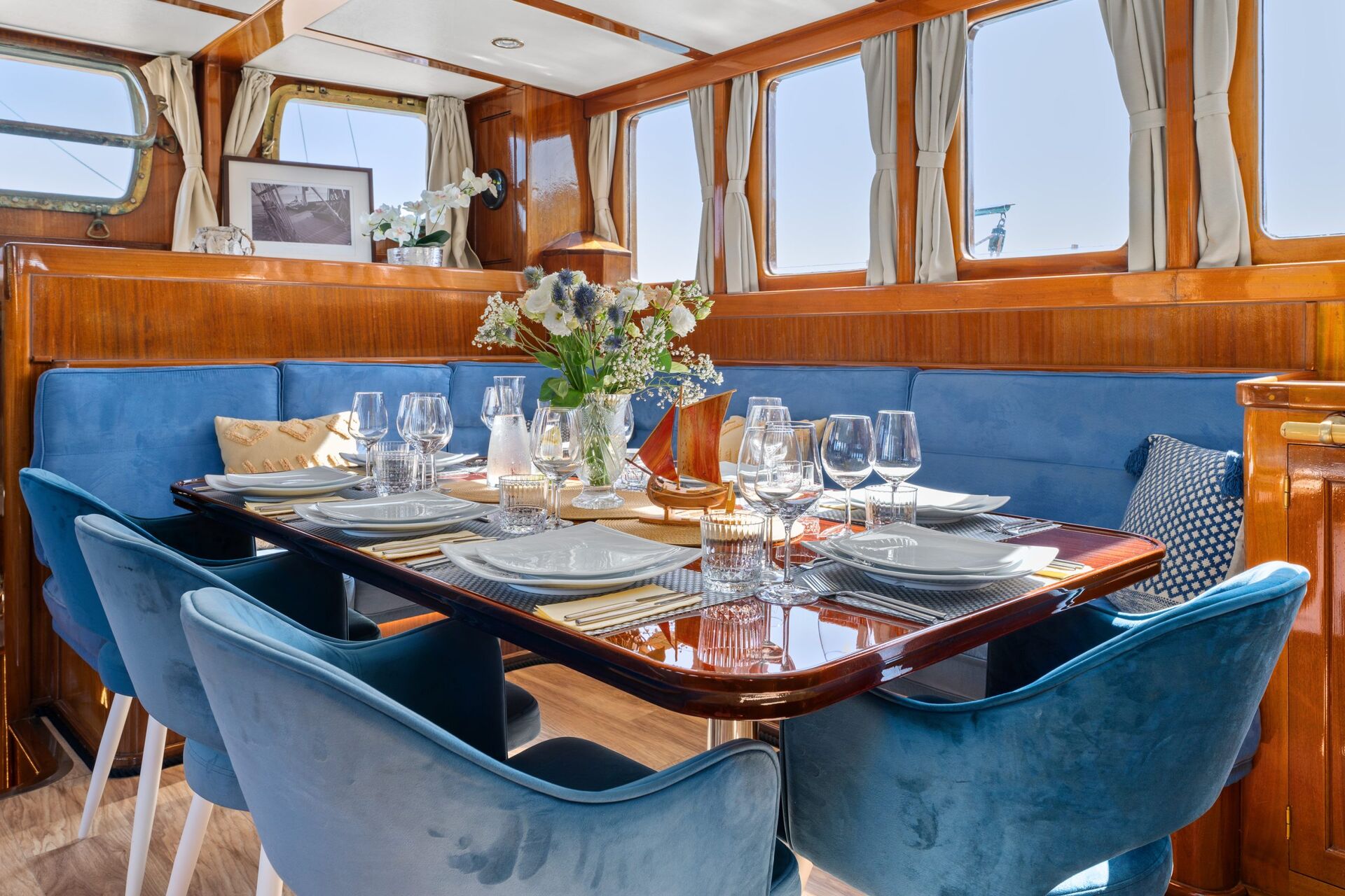 A dining area on a yacht features a polished wooden table set for six, with blue velvet chairs, elegant tableware, flowers, and large windows letting in natural light.