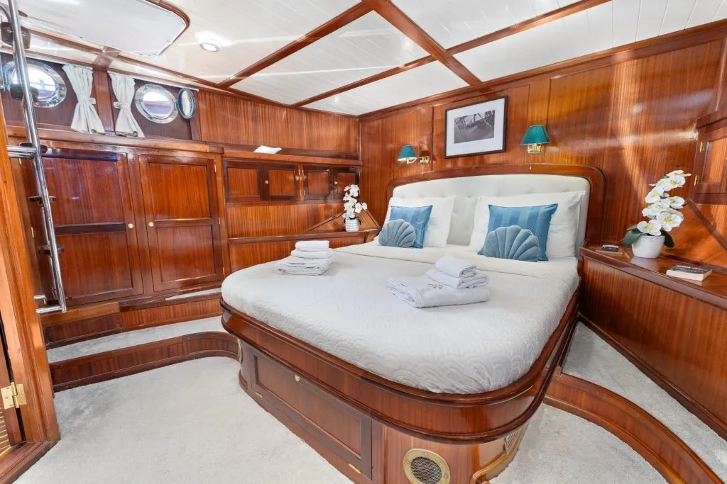 A cozy yacht cabin with polished wood paneling, a large bed with white bedding and blue accent pillows, folded towels, bedside lamps, porthole windows, and decorative white orchids.