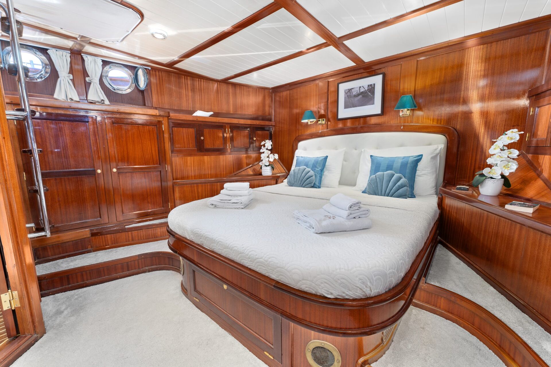 A cozy yacht cabin with polished wood paneling, a large bed with white bedding and blue accent pillows, folded towels, bedside lamps, porthole windows, and decorative white orchids.