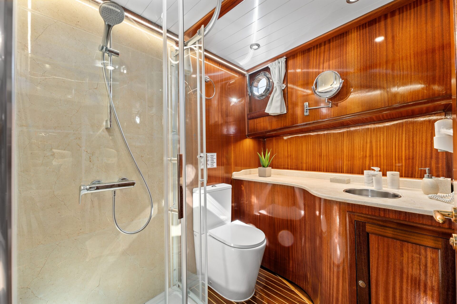 Luxurious yacht bathroom with polished wood walls, glass shower enclosure, rainfall shower, toilet, round porthole windows, white countertop with sink, soap dispensers, small potted plant, and elegant lighting.
