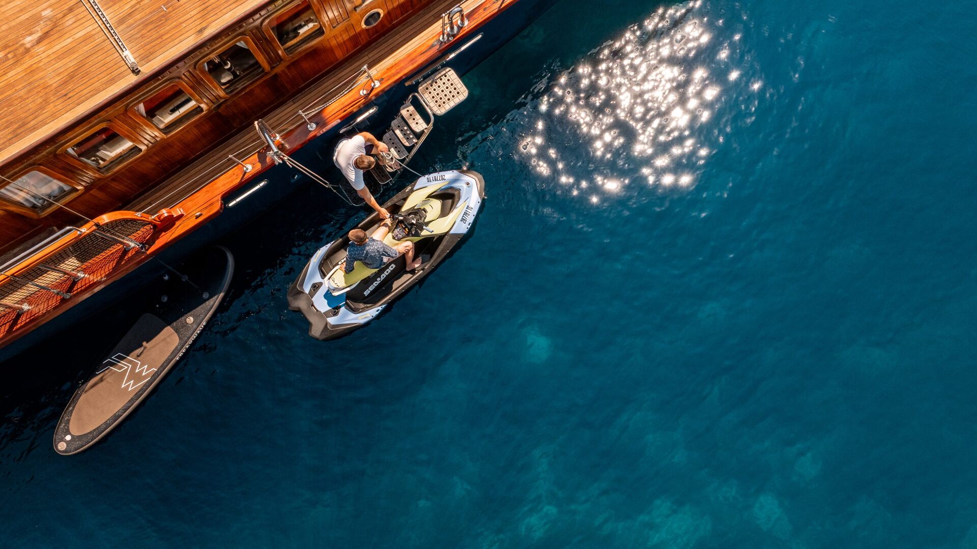 Aerial view of two people on a jet ski next to a wooden yacht in clear blue water, with sunlight reflecting on the surface and a paddleboard floating nearby.