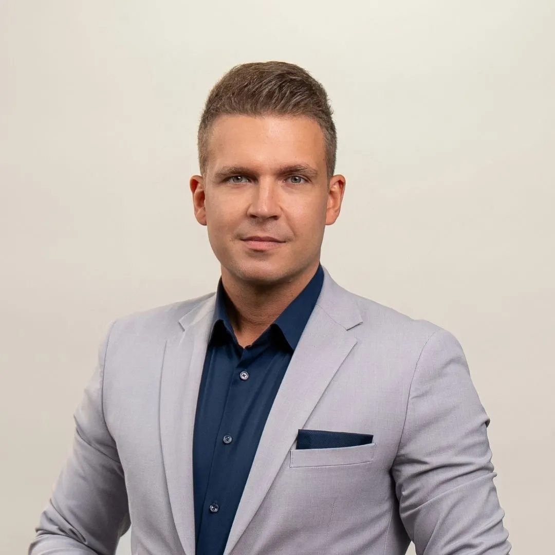 A man wearing a light gray suit jacket, dark navy shirt, and matching pocket square stands against a plain light background, looking directly at the camera with a neutral expression.