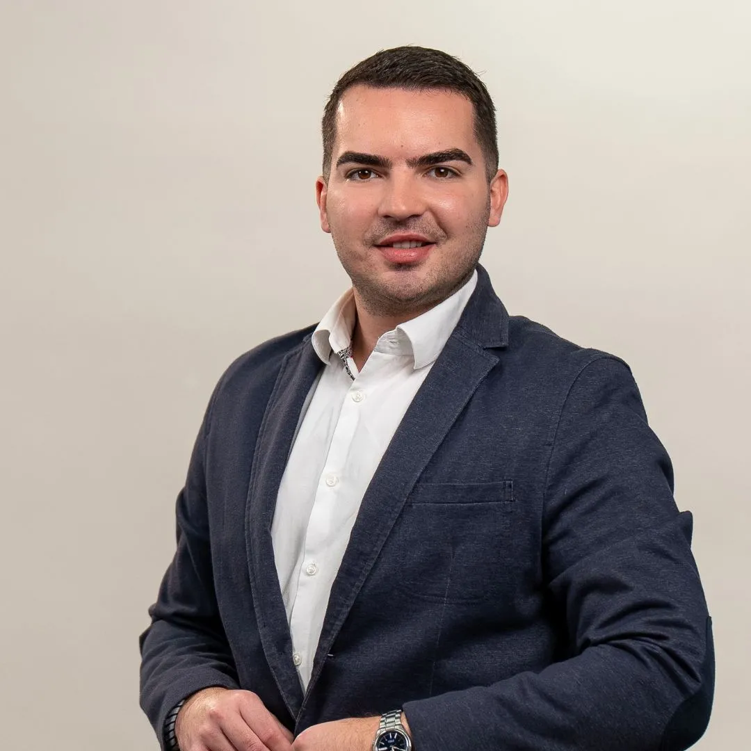 A man with short dark hair, wearing a white shirt and dark blazer, stands against a plain light background, looking at the camera with a slight smile and one hand adjusting his jacket.