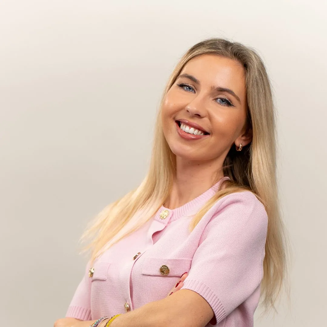 A woman with long blonde hair, wearing a light pink short-sleeved top, stands smiling confidently with her arms crossed against a plain, light background.