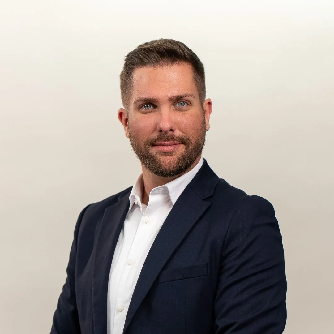 A man with short brown hair and a trimmed beard, wearing a navy blue suit jacket over a white dress shirt, poses confidently against a plain light background.