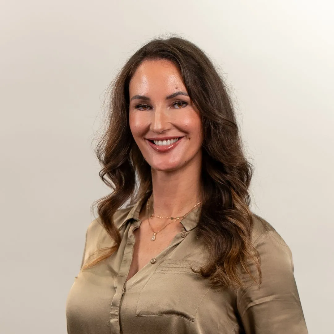 A woman with long, wavy brown hair wearing a light brown satin blouse and layered gold necklaces smiles at the camera against a plain, light background.