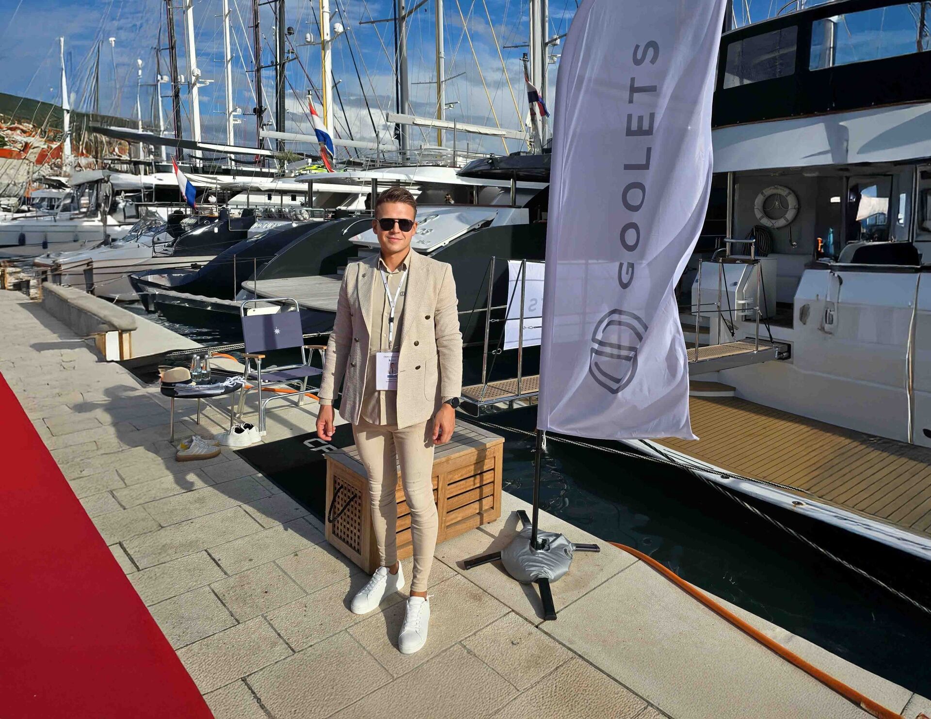 A man in a beige suit and sunglasses stands on a marina dock beside luxury yachts. A white flag with the GOOLETS logo is displayed next to him, and a red carpet runs along the left side of the dock.