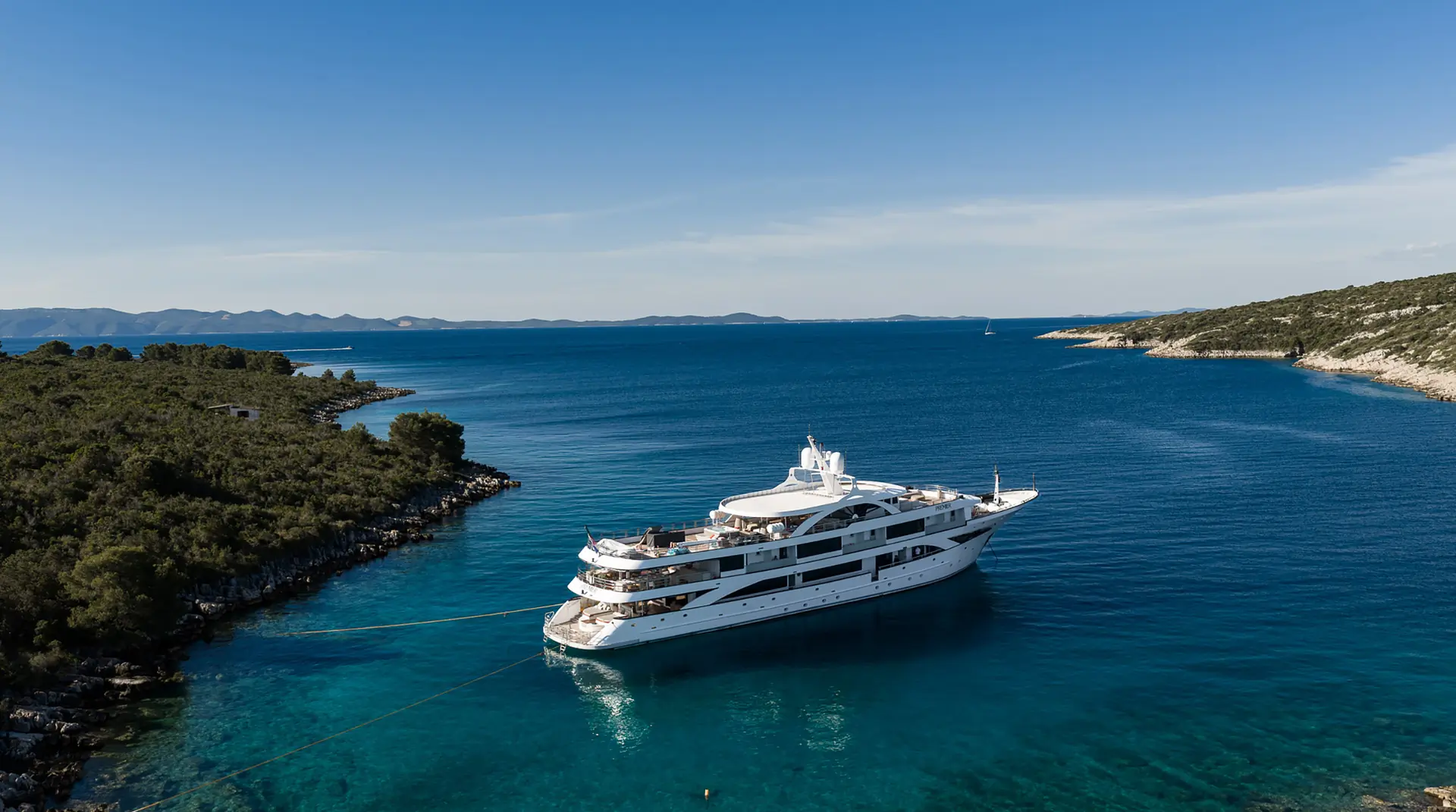A large white yacht anchored in clear blue water near a rocky, tree-covered coastline under a clear sky.