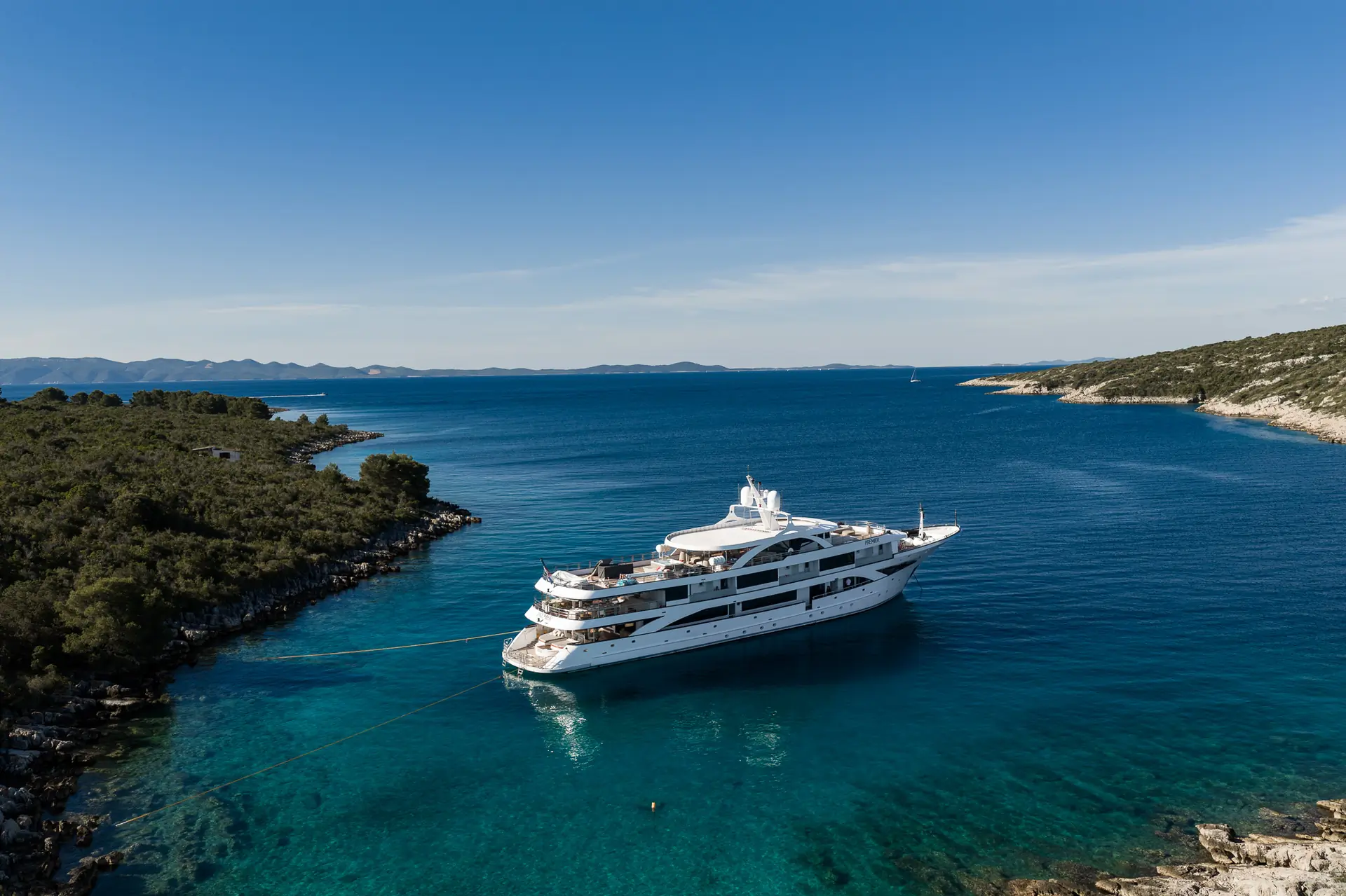A large white yacht is anchored in a calm, clear blue bay surrounded by green, tree-lined shores under a bright blue sky. The yacht is moored with ropes extending toward the land.