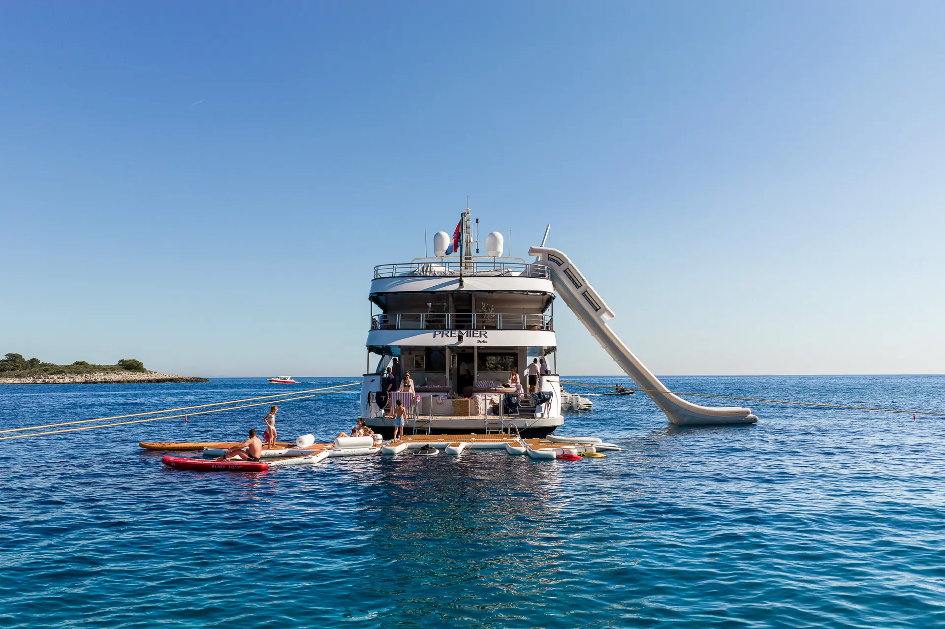 A large yacht anchored in blue water has a tall slide extending from its top deck into the sea. People are relaxing on paddleboards and kayaks around the yacht, with a small island visible in the distance.