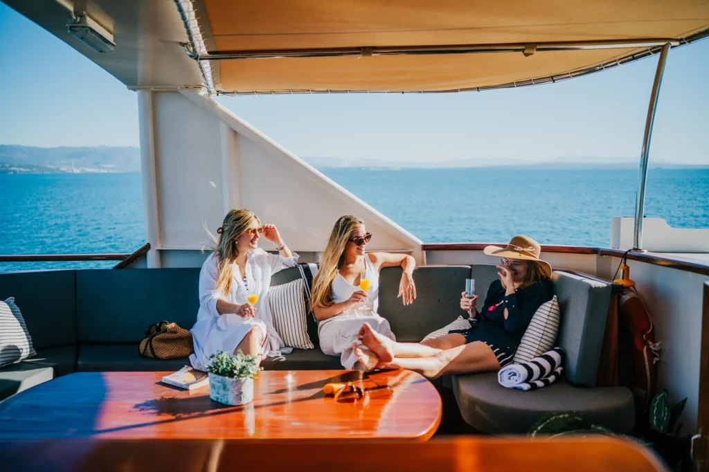 KORAB Three women relax on a yacht’s shaded deck, enjoying drinks and conversation. The sea and distant coastline are visible in the background under a clear blue sky.