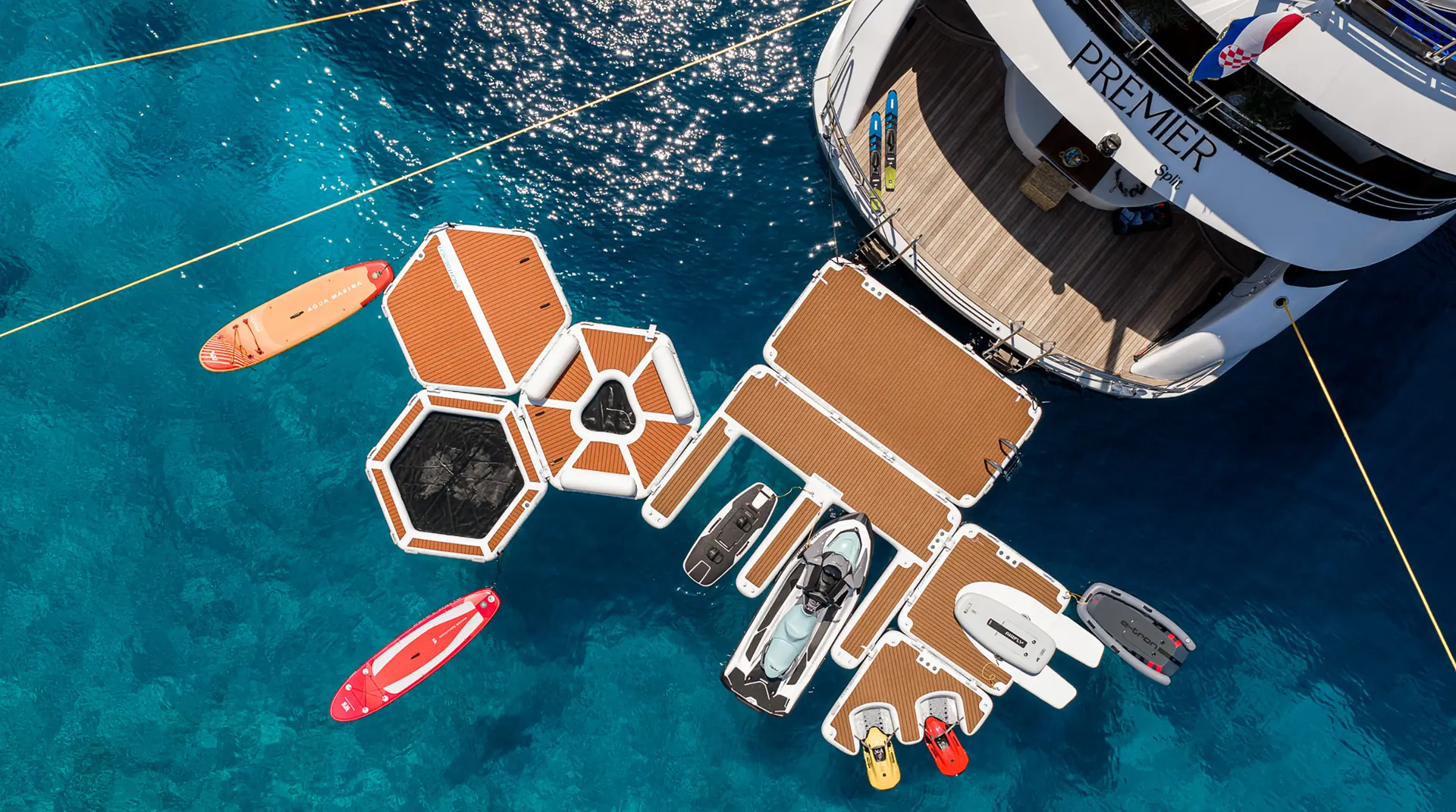 Aerial view of a yacht named Premier with modular floating docks attached, featuring paddleboards, jet skis, and small boats on clear blue water.