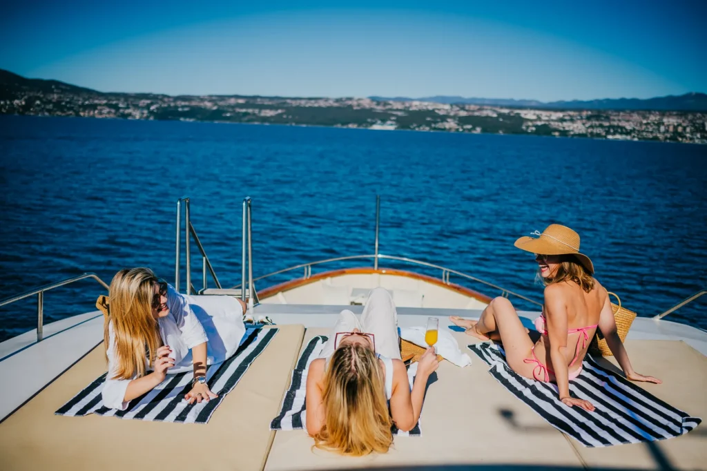 KORAB Three women relax on striped towels at the front of a boat, enjoying the sun. One drinks from a glass, another wears a sunhat, and the blue water and distant shoreline are visible in the background.