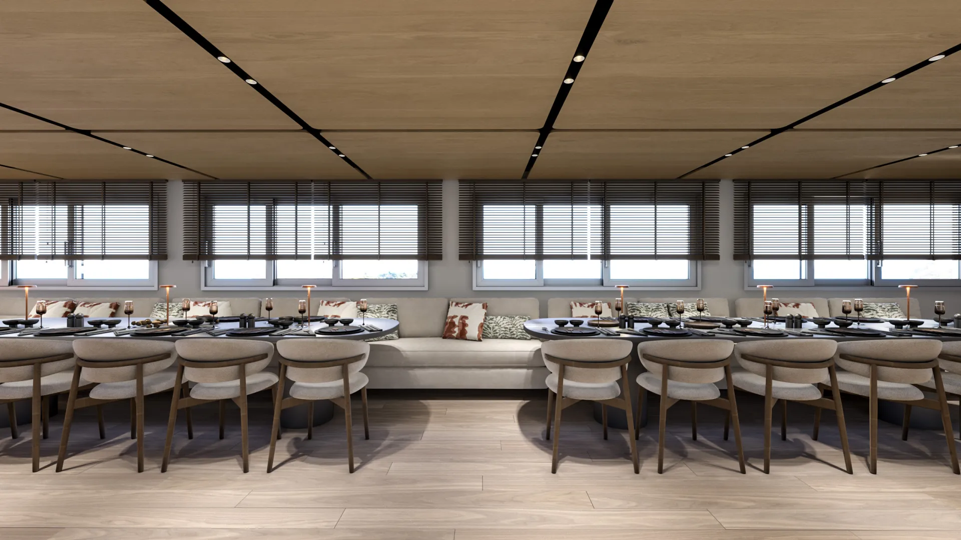 PREMIER A modern dining area with two long tables set for a meal, beige cushioned chairs, decorative pillows on a central bench, large windows with blinds, and wooden floors and ceiling panels.