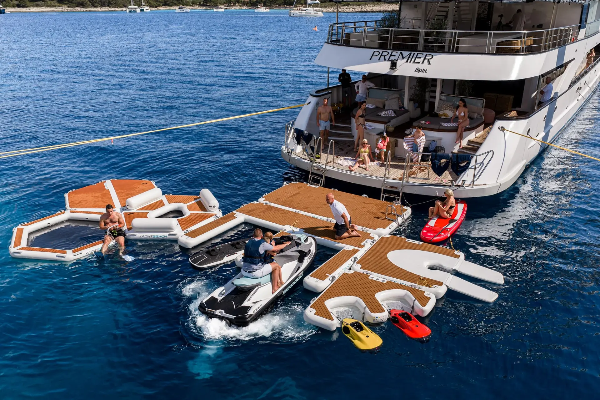 People relax and play on floating platforms, jet skis, and inflatable toys beside a large yacht anchored in clear blue water. Some lounge on the yacht’s deck while others interact on the floating dock and watercraft.