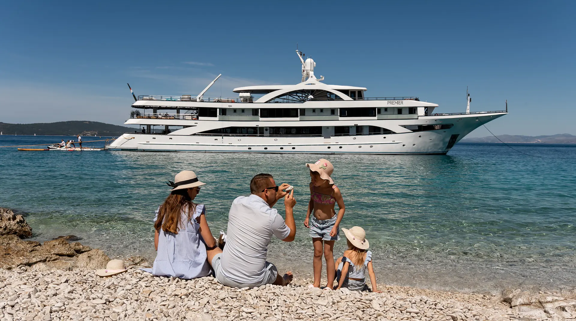 A family of four sits on a rocky beach facing clear blue water, with a large white yacht anchored nearby. The adults and children wear summer clothes and hats, enjoying a sunny day by the sea.