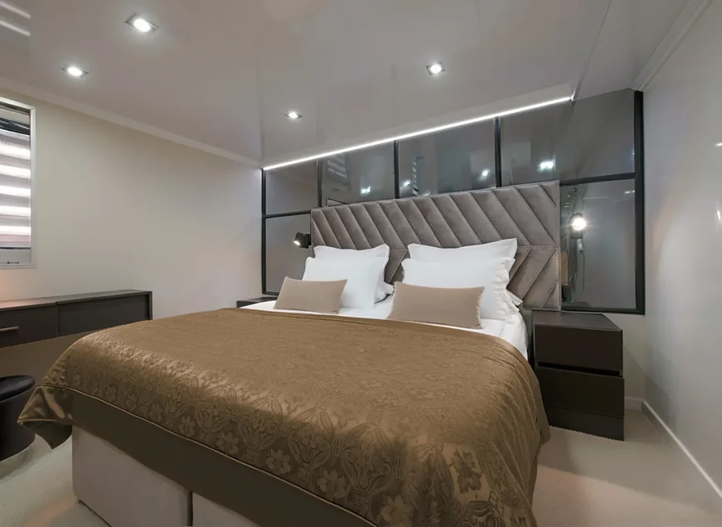 PREMIER Modern bedroom with a large bed featuring a brown patterned bedspread, white and beige pillows, a padded headboard, glass wall accents, bedside tables, and built-in ceiling lights. The decor is sleek and minimalist.