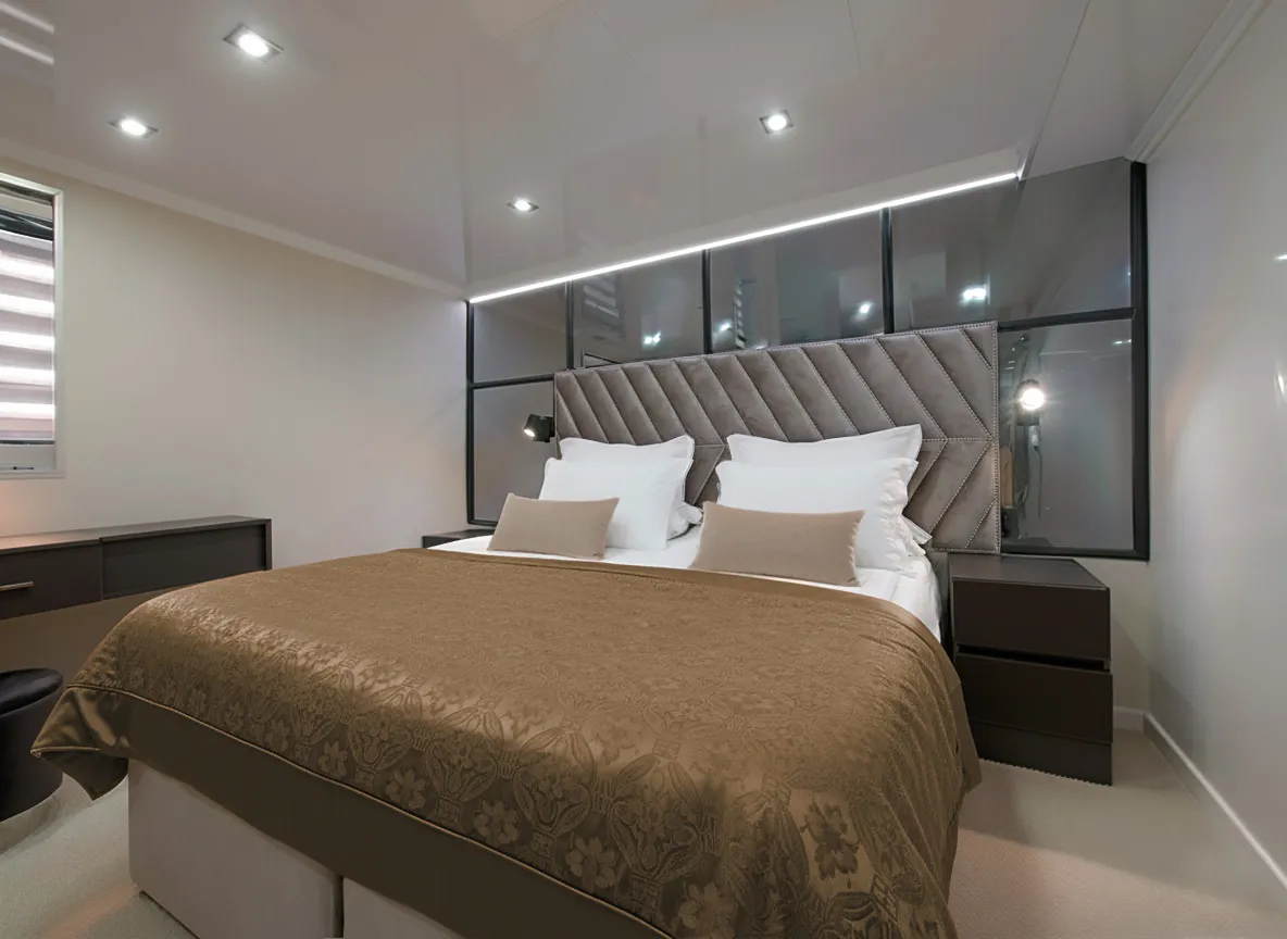 PREMIER Modern bedroom with a large bed covered in a brown patterned bedspread, white pillows, two bedside tables with lamps, and a padded headboard against a sleek, mirrored wall. Room is well-lit and minimalist.