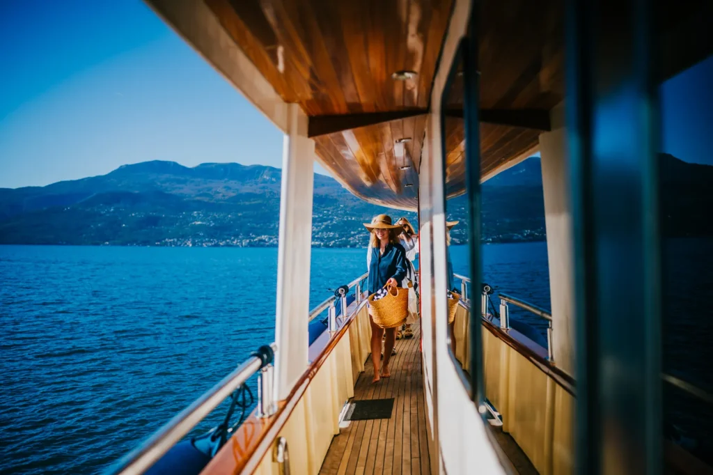 KORAB A person wearing a hat and carrying a woven basket walks along the deck of a boat, with calm blue water and distant mountains visible in the background under a clear sky.