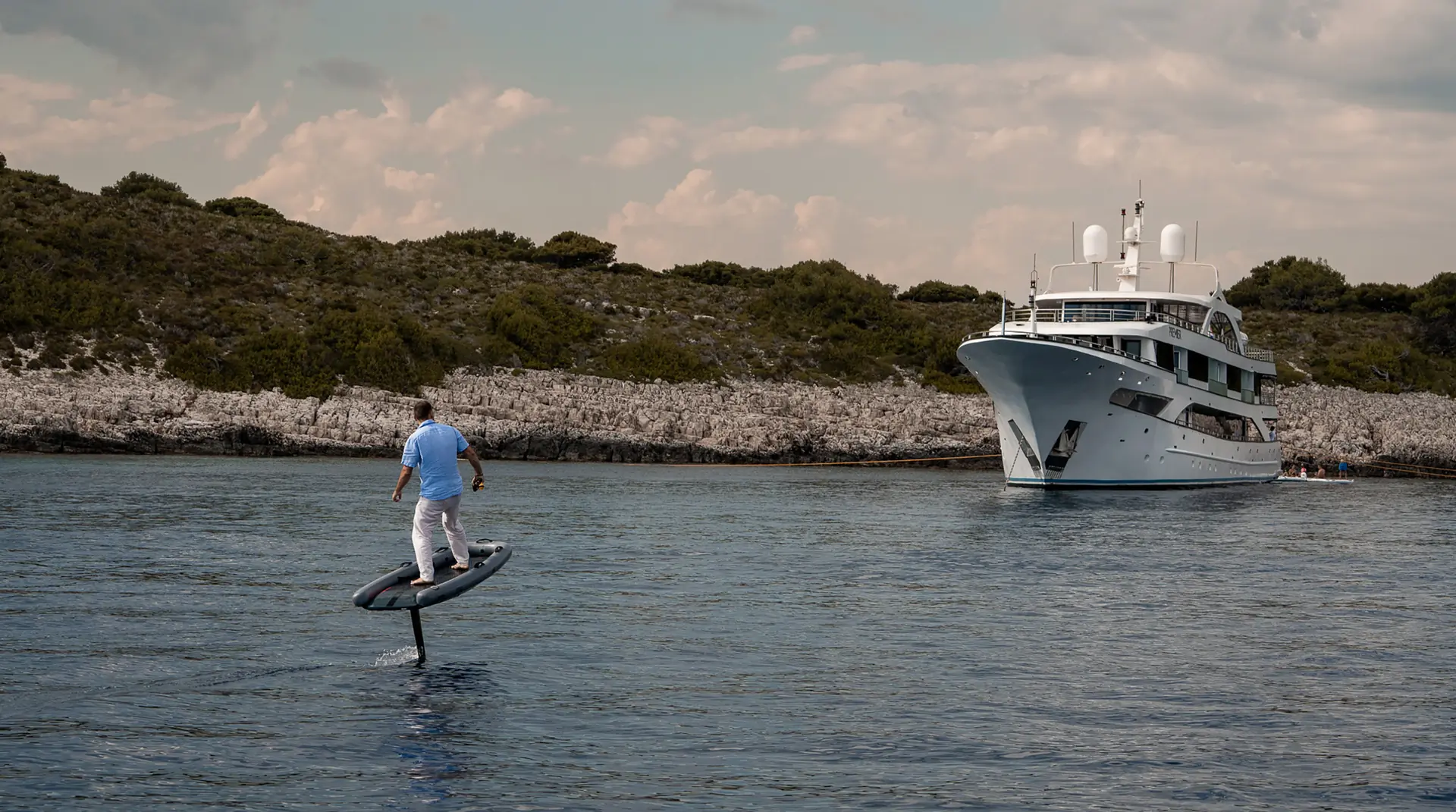 A person in light clothing rides an electric hydrofoil surfboard on calm water near rocky shore, with a large white yacht anchored in the background under a partly cloudy sky.