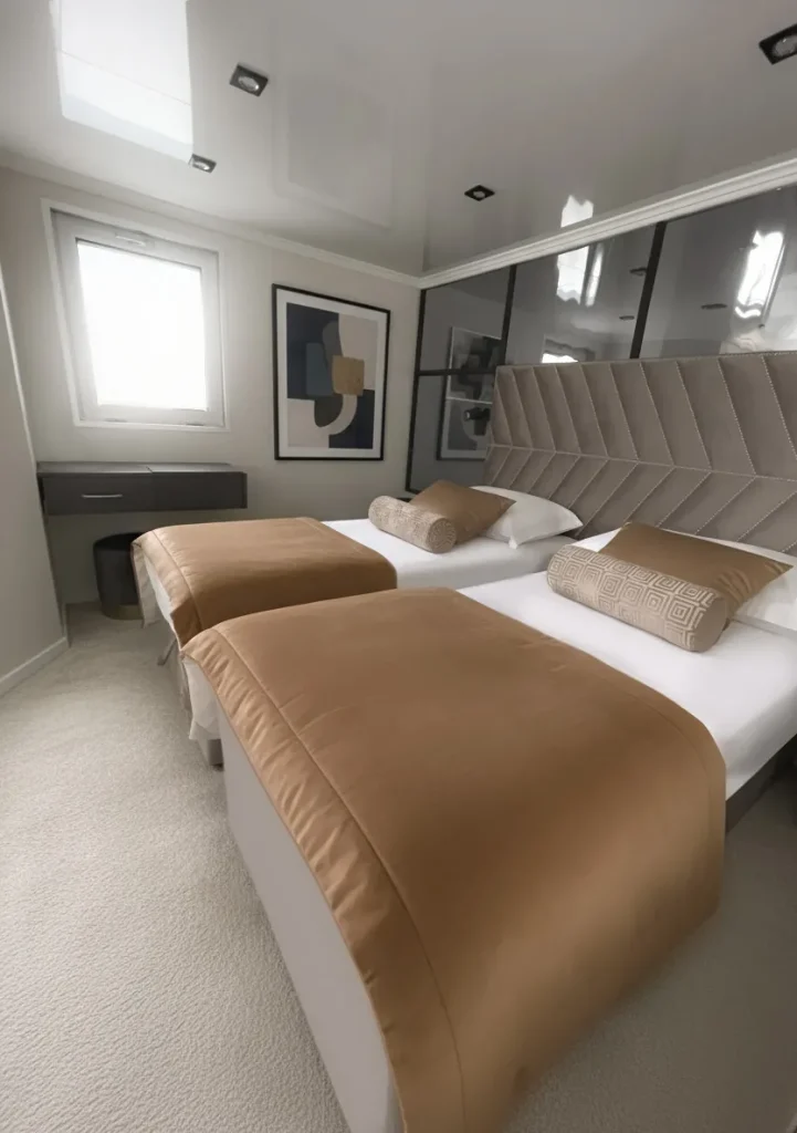 PREMIER A modern bedroom with two single beds, tan bedding, geometric padded headboard, abstract art on the wall, light carpet, a small desk under a window, and neutral-colored decor.