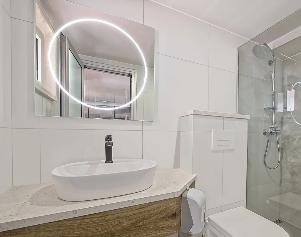 PREMIER Modern bathroom with a round, backlit mirror above a white sink and black faucet, a glass shower enclosure with rain showerhead, and light-colored tiled walls.