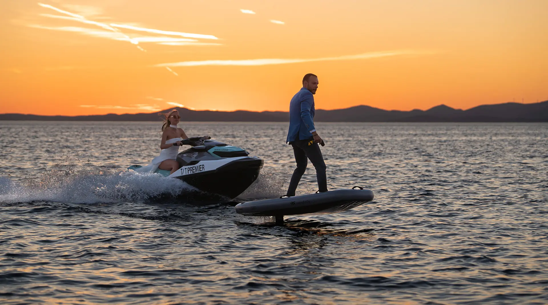 A man in a suit rides an electric surfboard on calm water at sunset, while a woman follows behind on a jet ski. Hills are visible in the background beneath an orange sky.