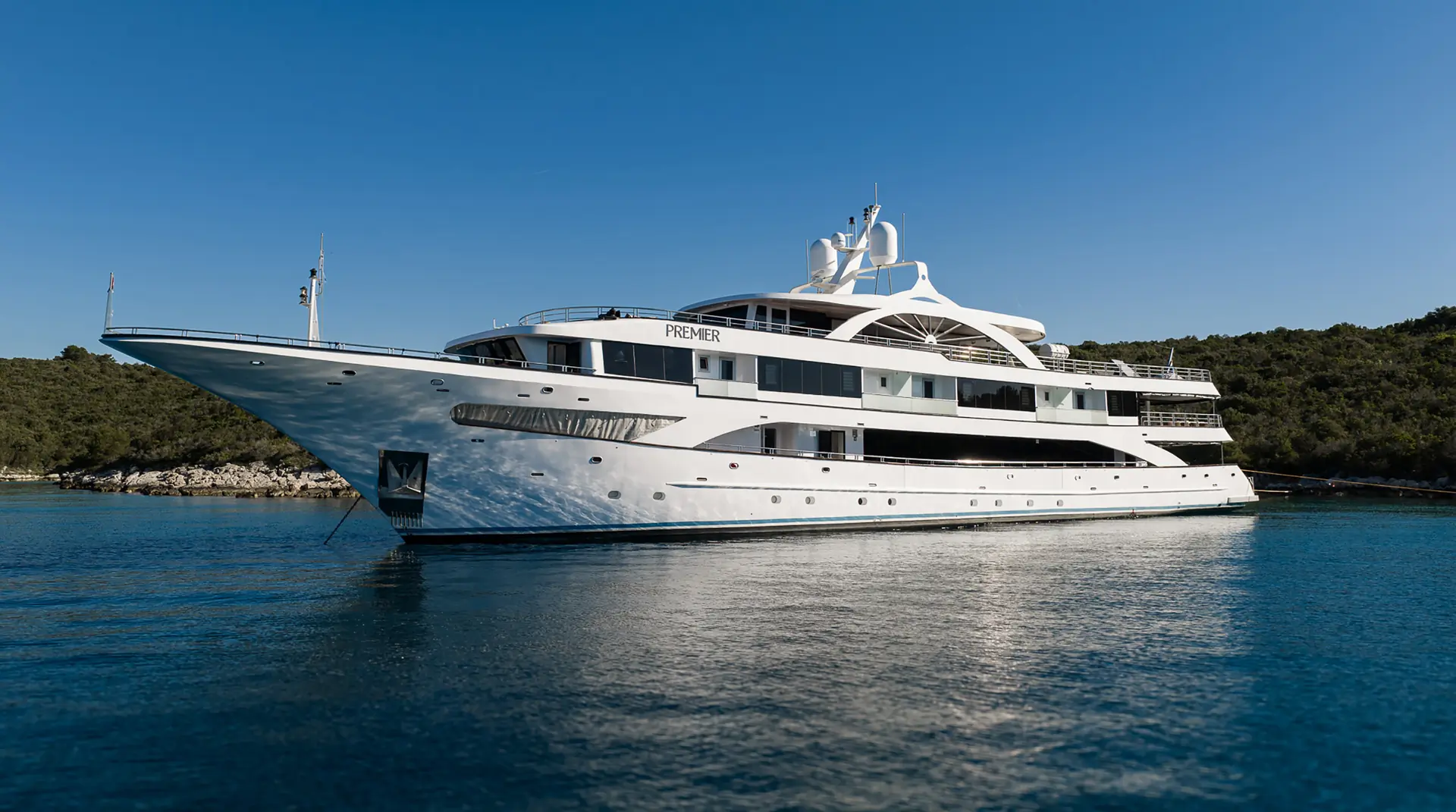 A large, luxurious white yacht named Premier is anchored on calm blue water, with green trees and clear blue sky in the background.