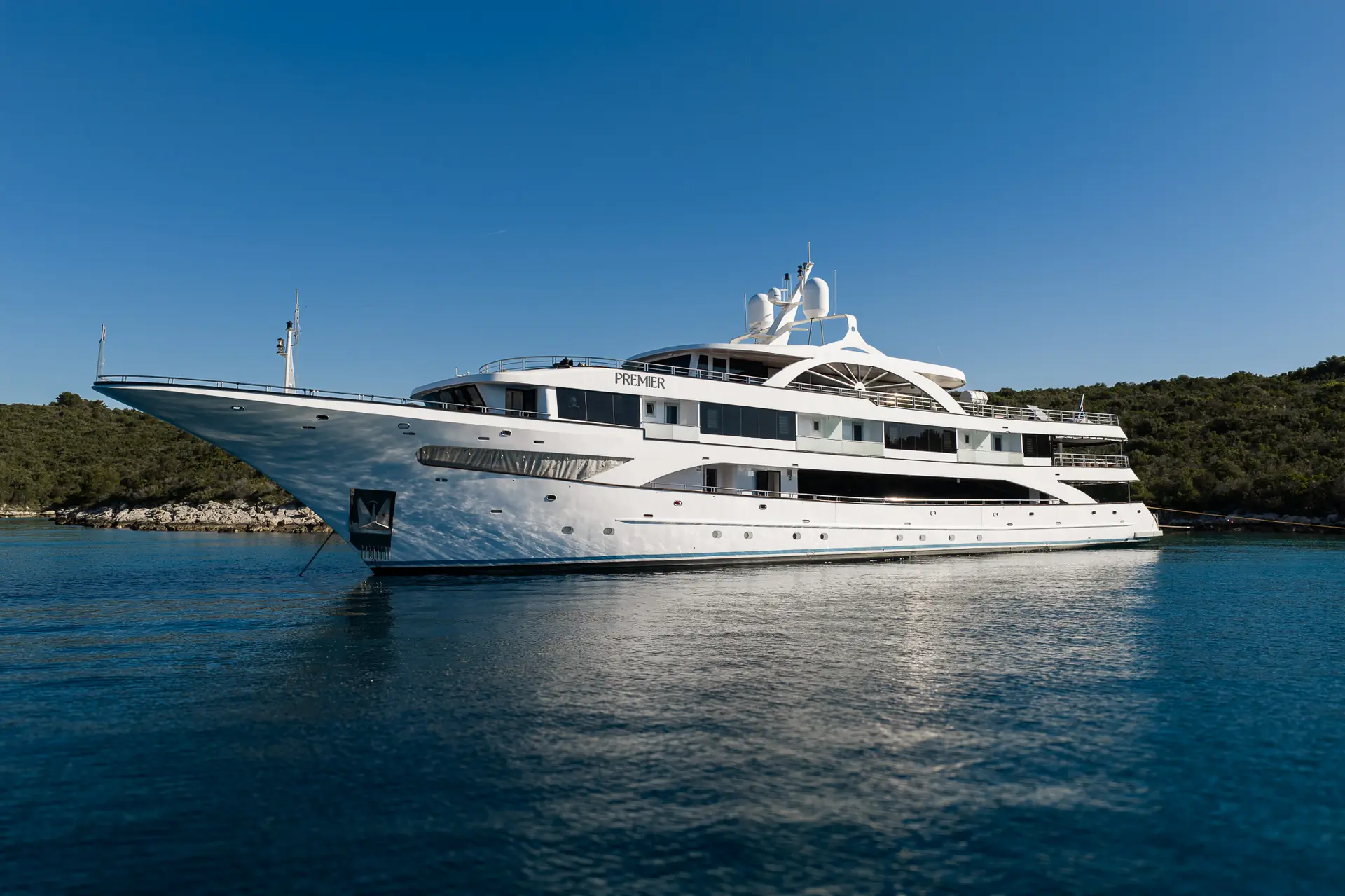 A large white luxury yacht named Premier floats on calm blue water near a green, tree-covered shoreline under a clear blue sky.