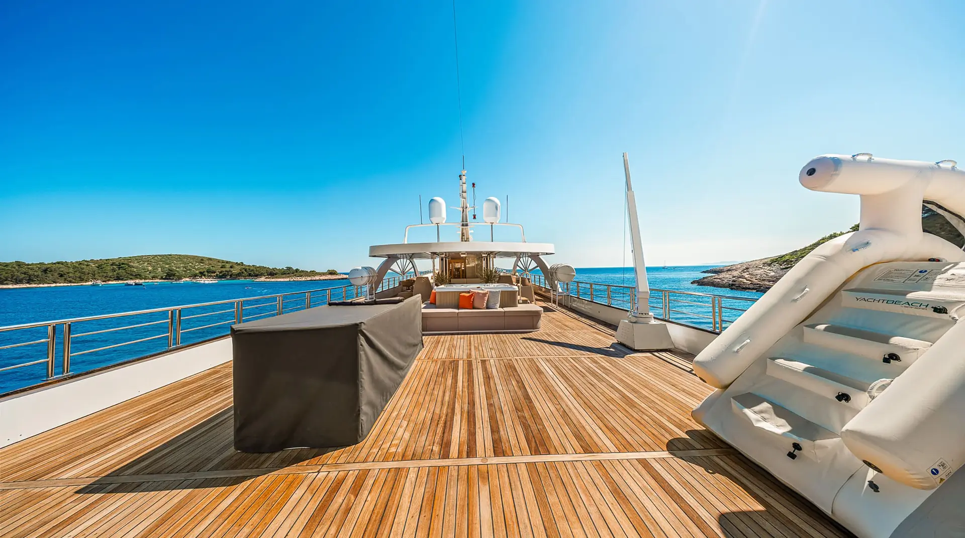 The sun deck of a luxury yacht with wooden flooring, lounge seating, and an inflatable water slide, surrounded by calm blue sea and distant green islands under a clear blue sky.