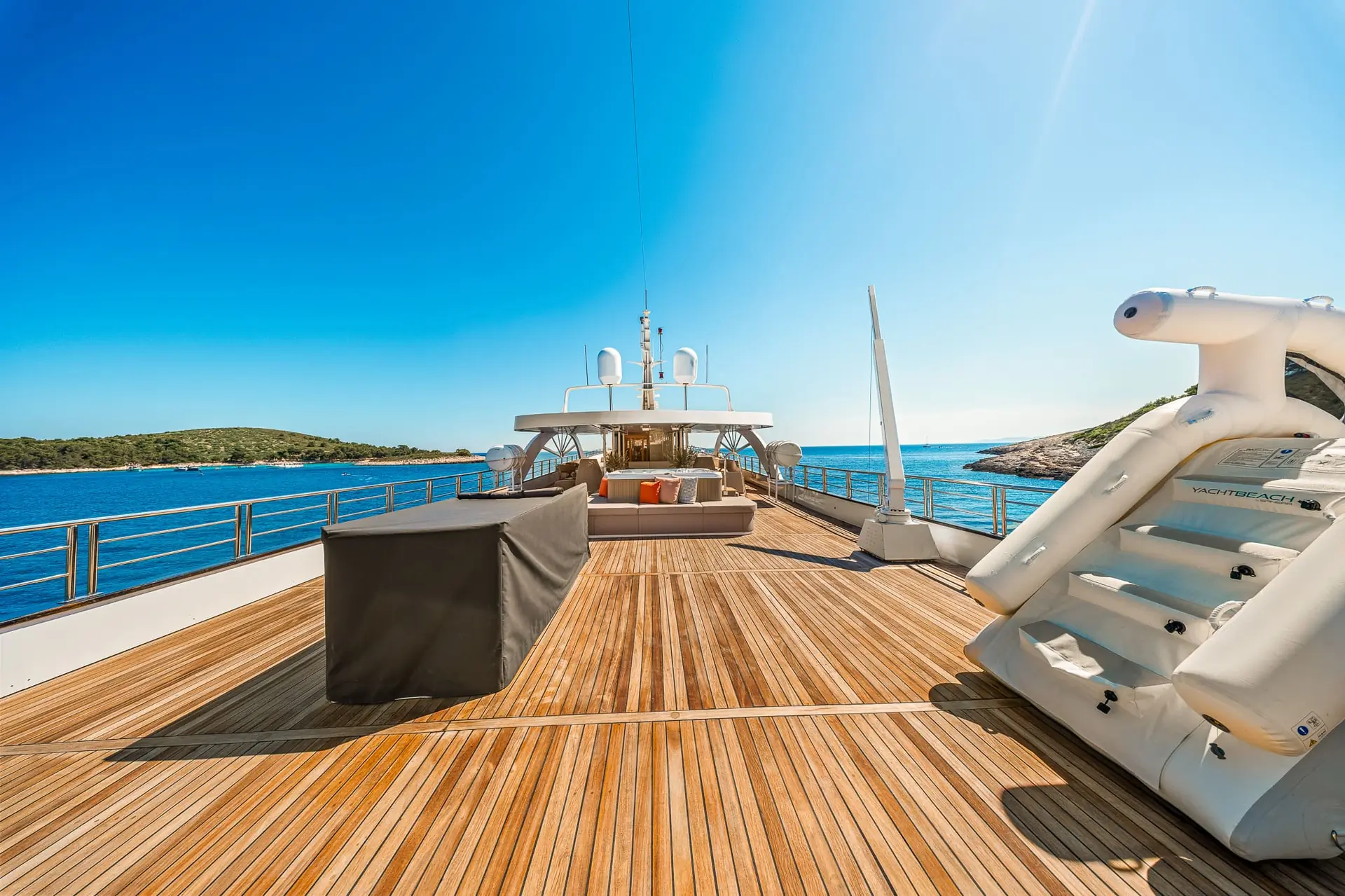 Spacious wooden deck of a luxury yacht with outdoor seating, surrounded by clear blue sea and distant green islands under a bright, sunny sky.