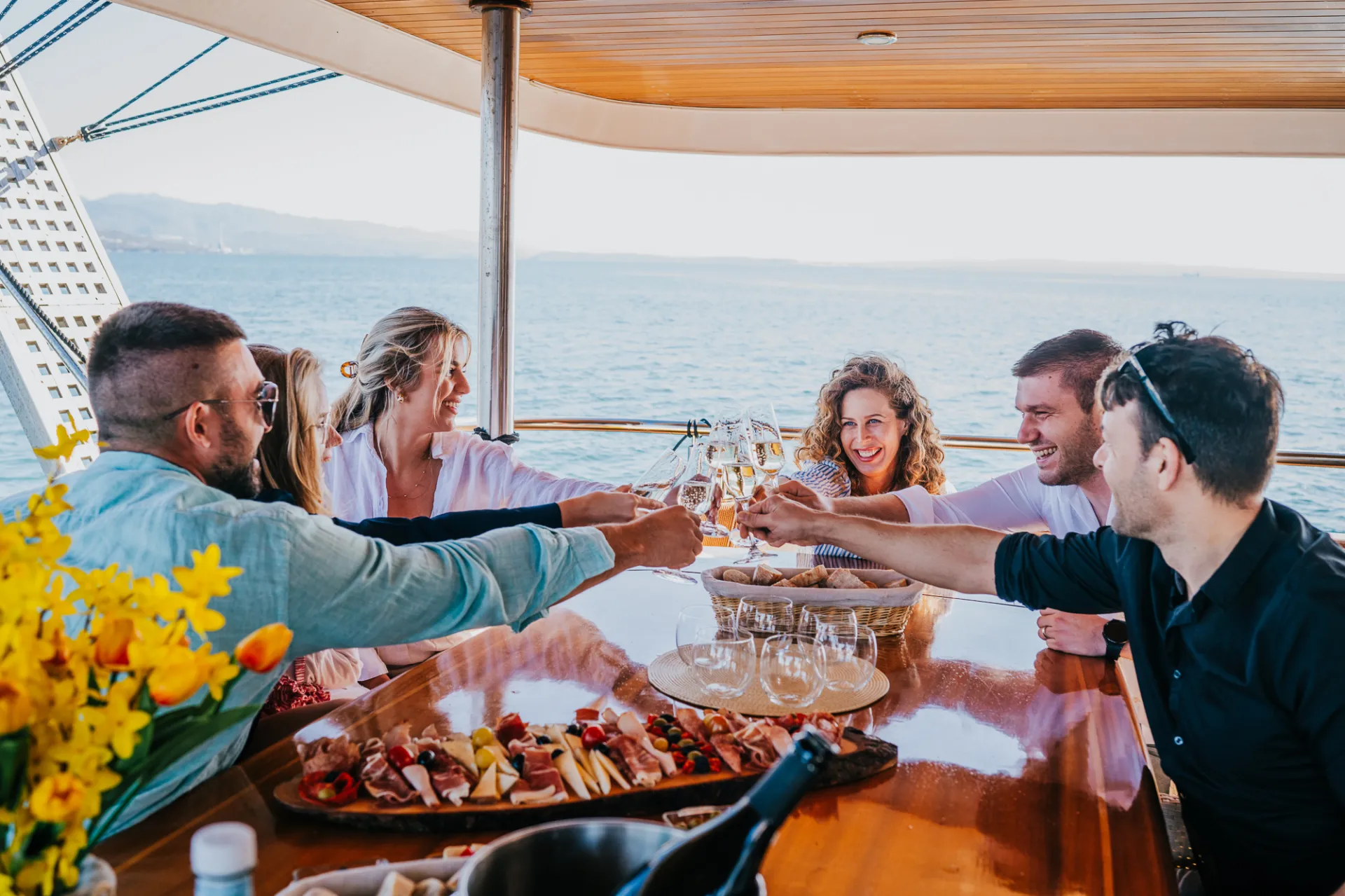 KORAB Six people sit around a table on a boat, smiling and clinking glasses in a toast. The table has plates of food and flowers, and the ocean is visible in the background under a clear sky.
