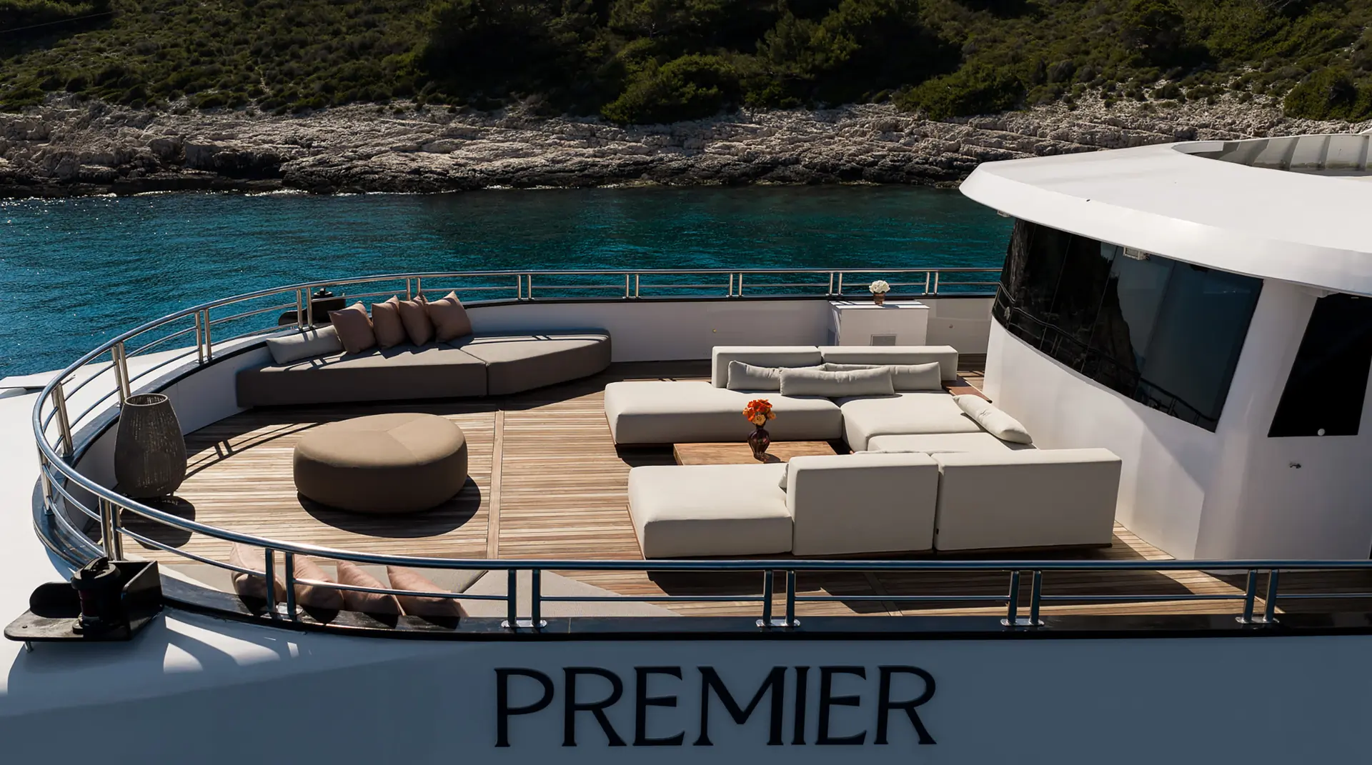 A luxury yacht deck with modern white sofas, a round ottoman, and a table with flowers, overlooks clear turquoise water and a rocky, tree-covered shoreline. The word PREMIER is visible on the yachts side.