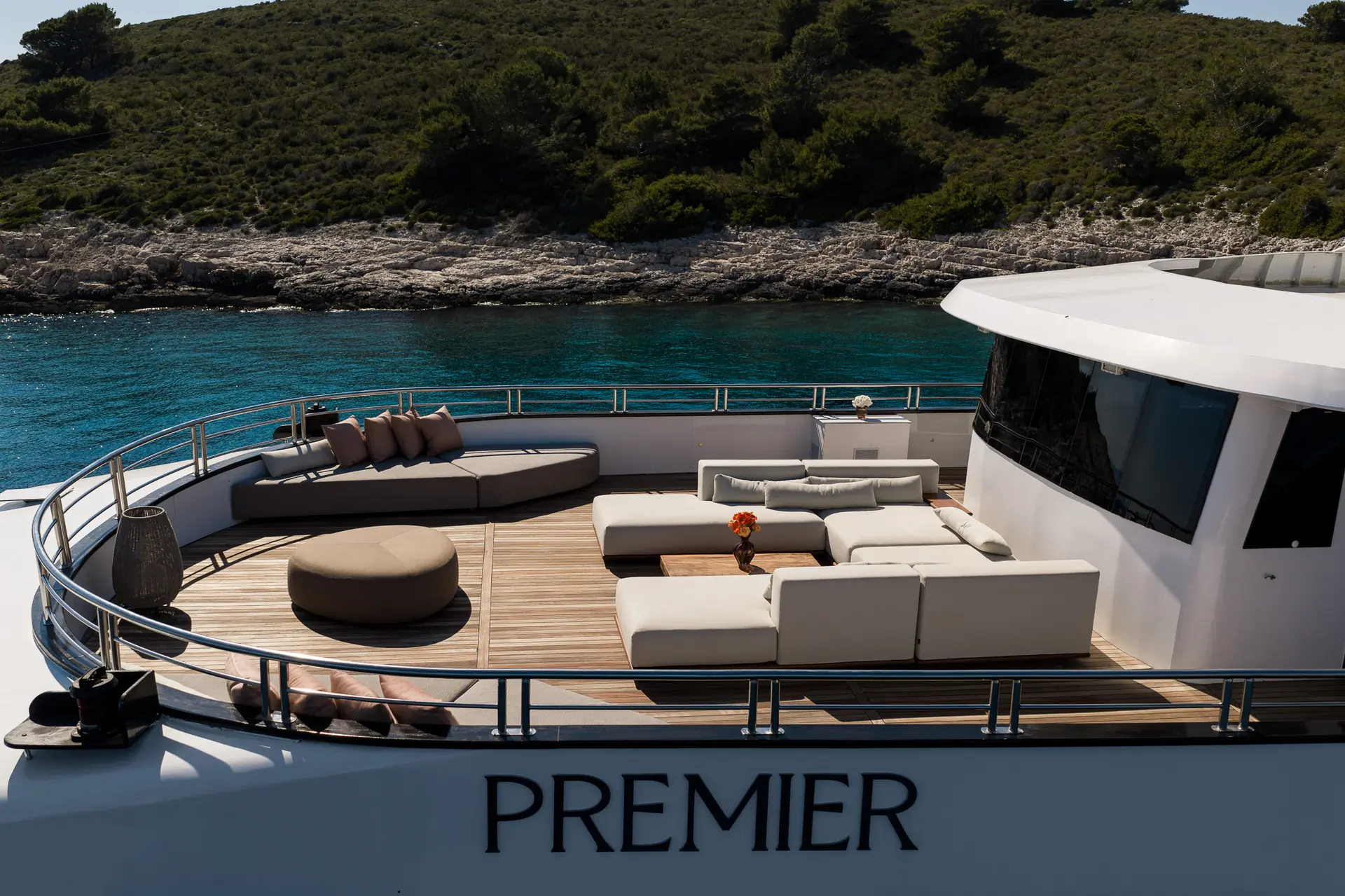 The deck of a luxury yacht features modern white lounge seating and a round ottoman, with clear turquoise water and a green, rocky shoreline in the background. The word PREMIER is visible on the yacht.
