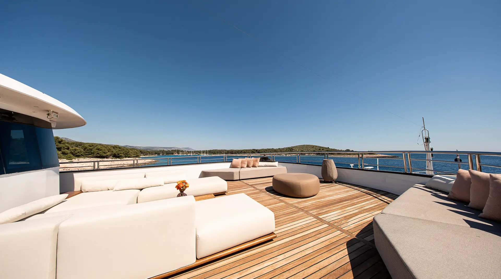 Spacious yacht deck with modern white sofas and beige cushions, wooden flooring, and ocean views under a clear blue sky. Distant land and calm water surround the stylish outdoor seating area.