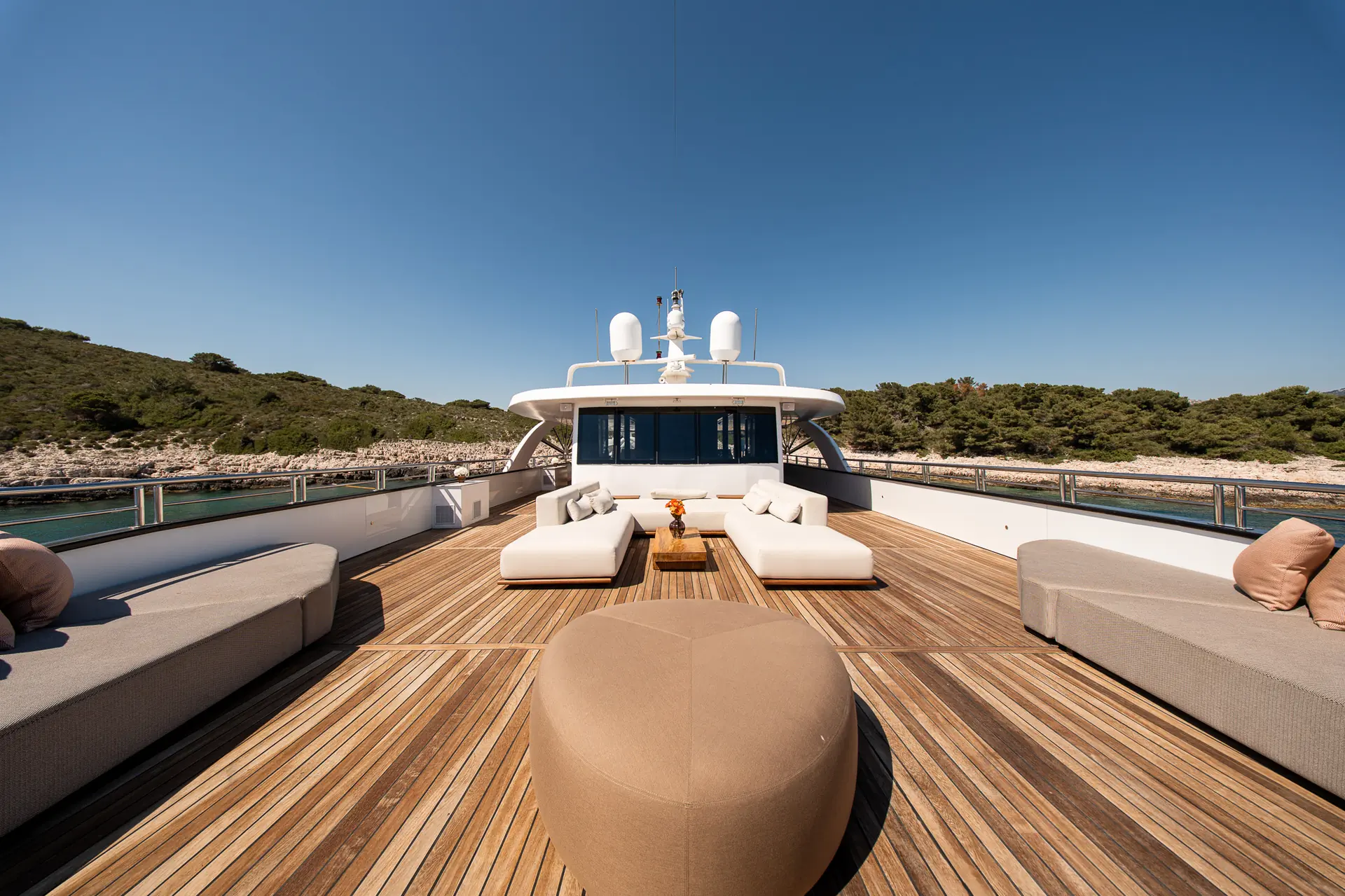 The image shows the deck of a luxury yacht with cushioned lounge seating and a central ottoman, surrounded by calm water and lush green trees under a clear blue sky.