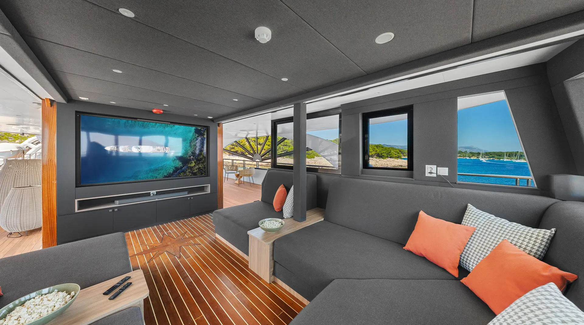 A modern yacht lounge with dark gray sofas, orange and patterned pillows, a large TV showing an underwater scene, wood flooring, and large windows revealing blue water and green islands outside.