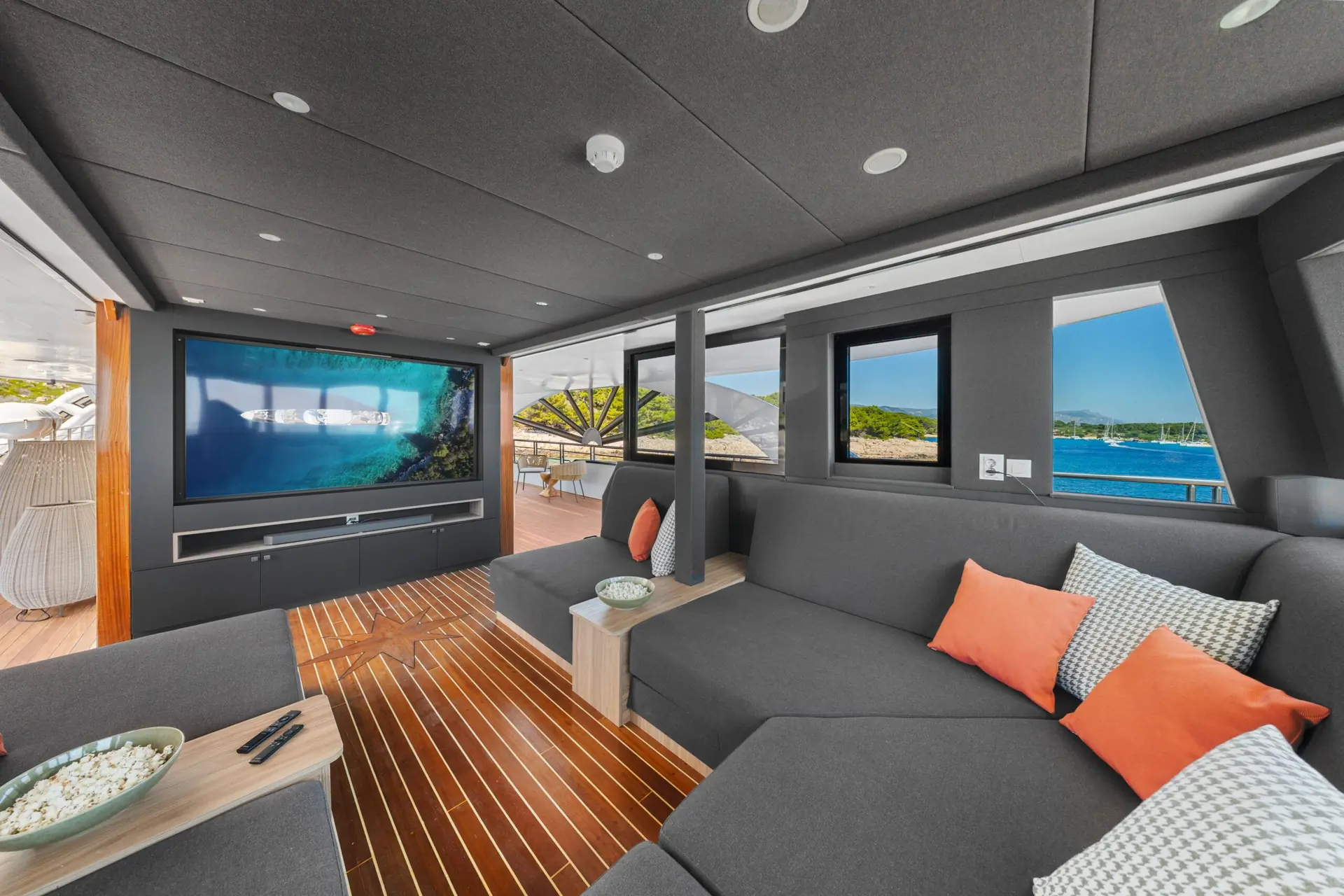 Modern yacht interior with dark gray sofas, orange and patterned pillows, wooden flooring, a large TV showing an ocean scene, and windows revealing a view of water and distant greenery. A bowl of popcorn sits on the table.