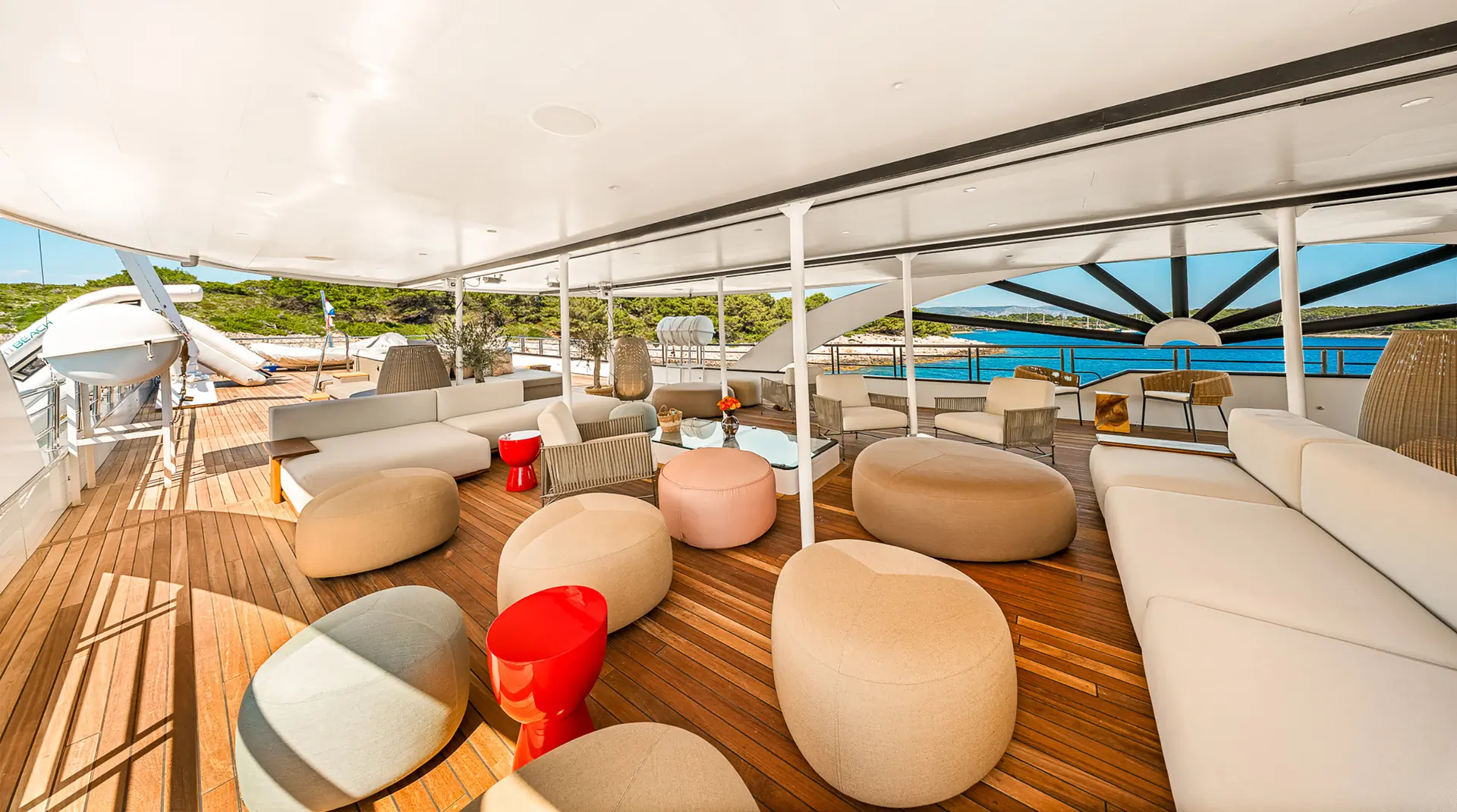 Luxurious yacht deck with modern, beige and tan cushioned seating, round stools, red side tables, and wooden flooring, overlooking calm blue water and a lush green shoreline in the background.