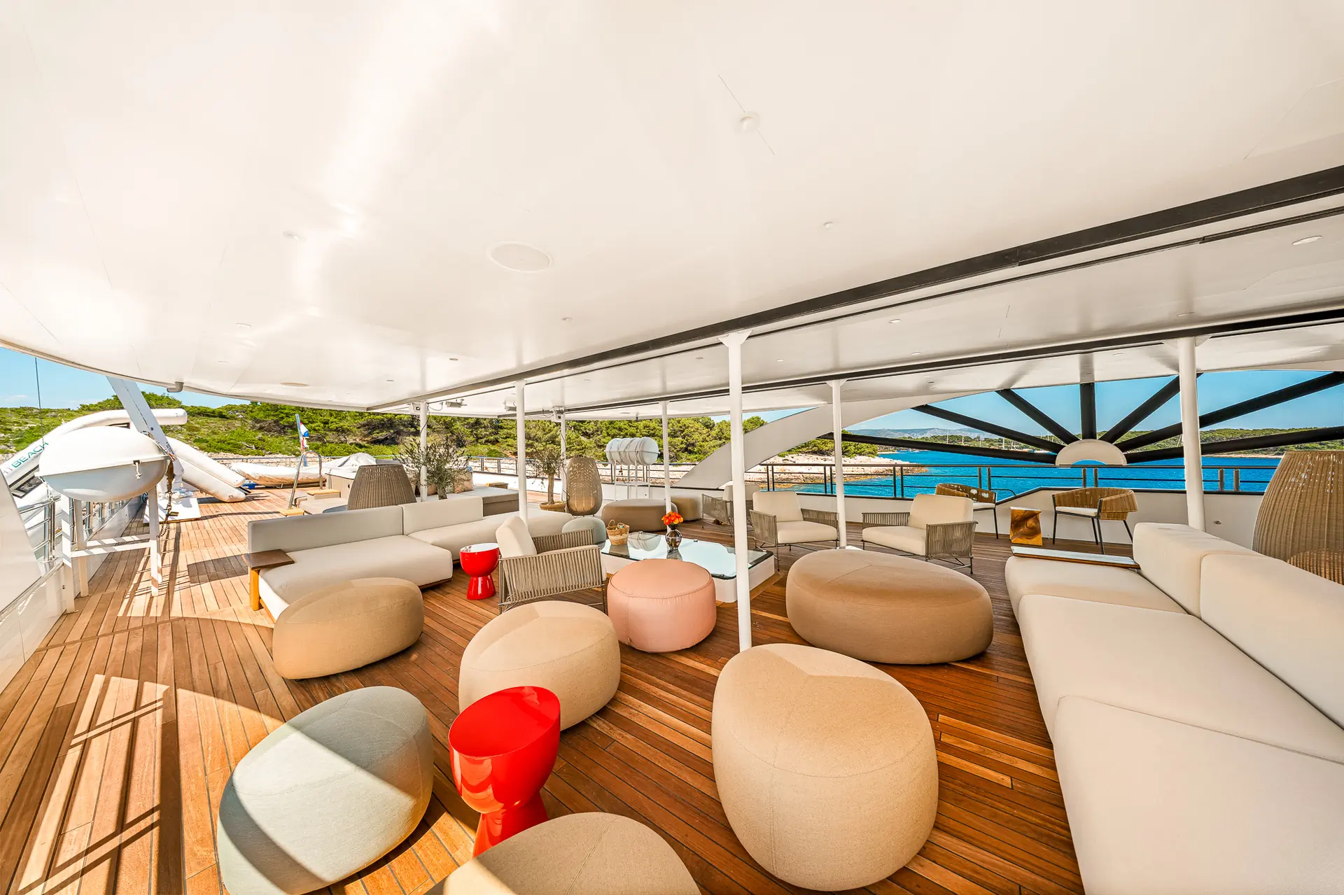 Spacious yacht deck with modern beige and tan seating, round ottomans, red side tables, and views of blue water and greenery under a white canopy. Relaxing, stylish outdoor lounge area.