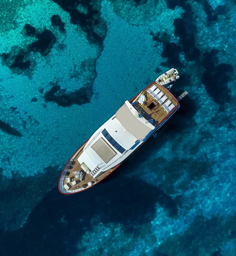 KORAB Aerial view of a white yacht floating on clear, deep blue water with lighter turquoise patches beneath the surface, indicating shallow areas or coral reefs.