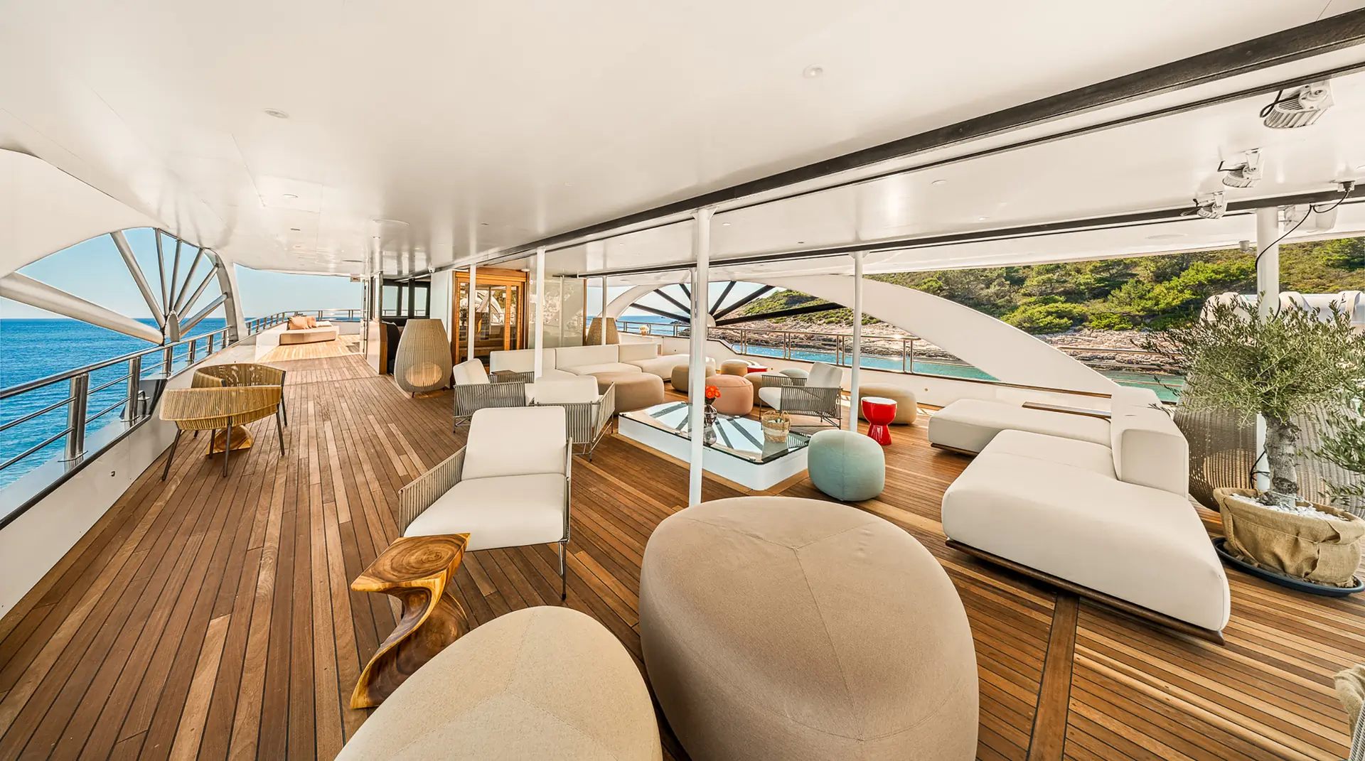 A spacious yacht deck with wooden flooring, modern beige and white seating, glass railings, and ocean and greenery views in the background under a white canopy.