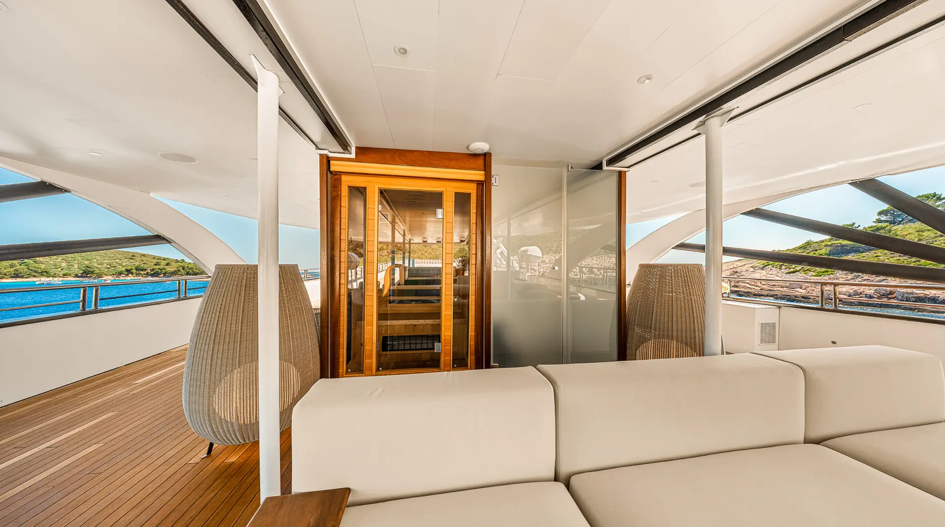 A modern yacht deck with a white cushioned sofa, wooden flooring, glass doors leading to an interior room, decorative lamps, and views of the ocean and coastal cliffs through large open windows.