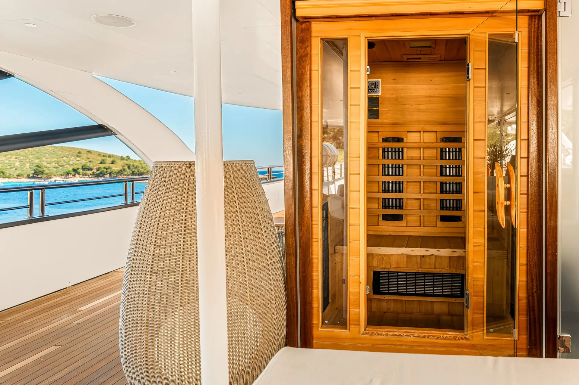 A wooden sauna with glass doors is located on the deck of a yacht, overlooking the sea and a distant green shoreline, with modern outdoor seating nearby.