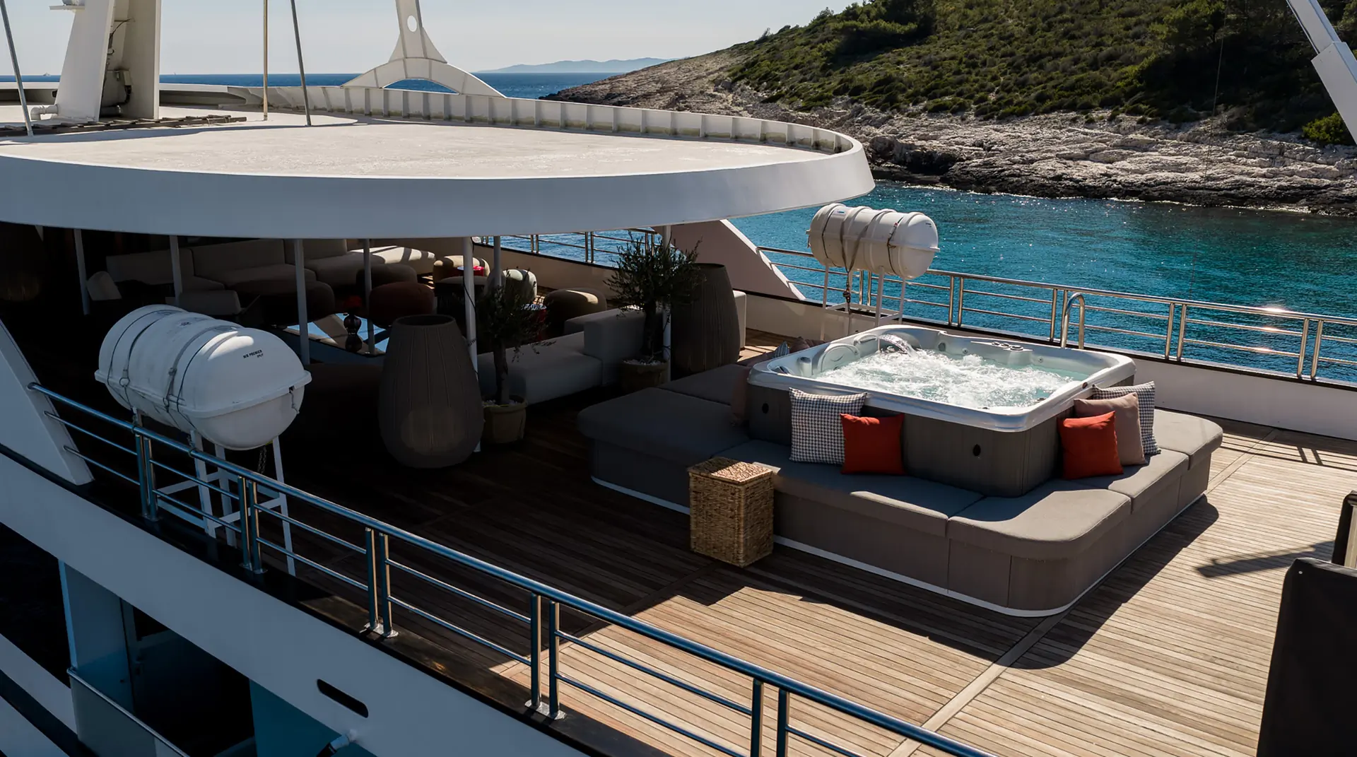 A luxurious yacht deck features a hot tub with bubbling water, surrounded by gray loungers and red cushions, overlooking clear blue sea and a rocky, green shoreline in the background.