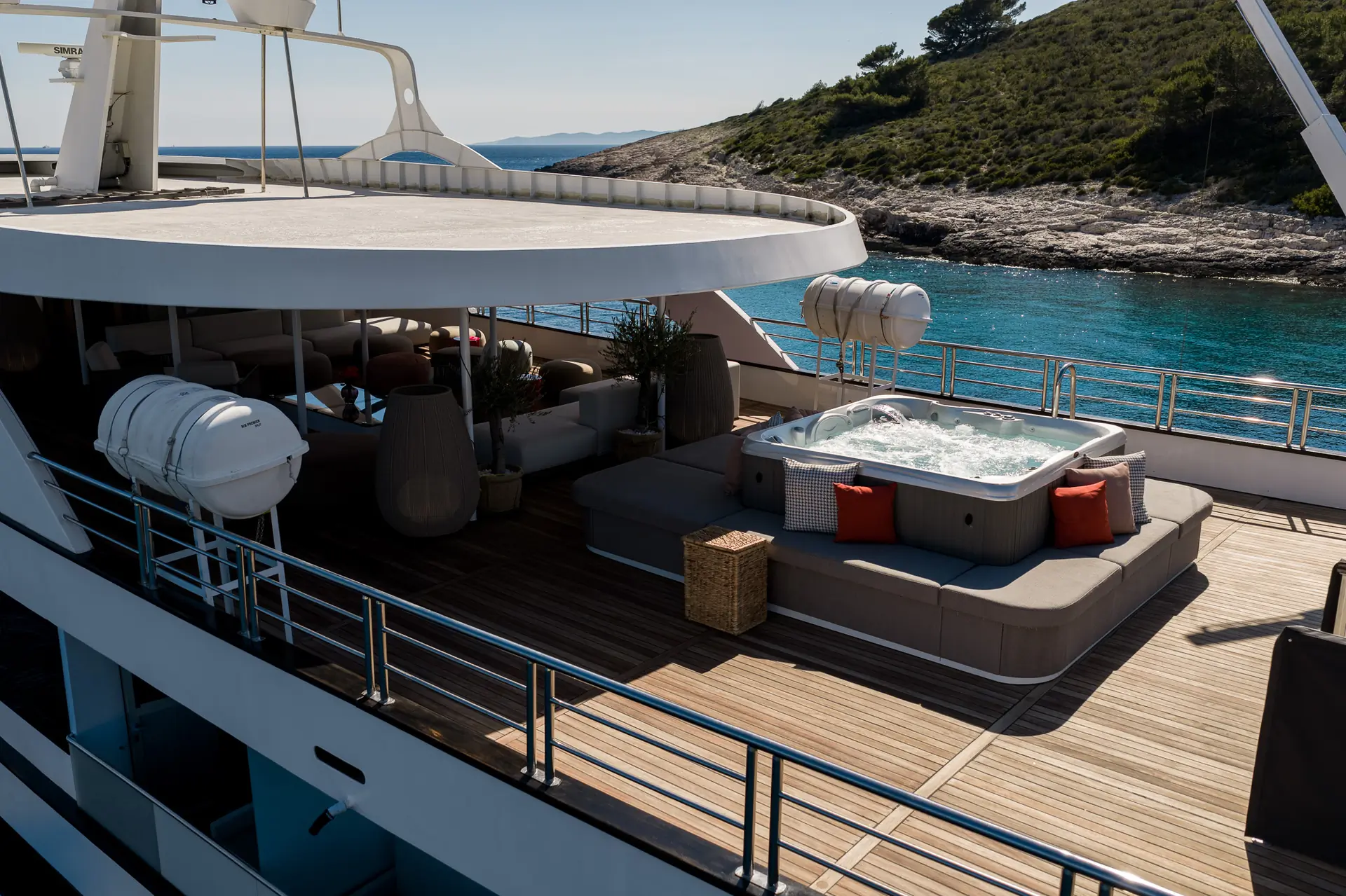 A luxury yacht deck featuring a hot tub with bubbling water, surrounded by cushioned seating and red pillows, overlooking clear blue water and a rocky, green coastline under a sunny sky.