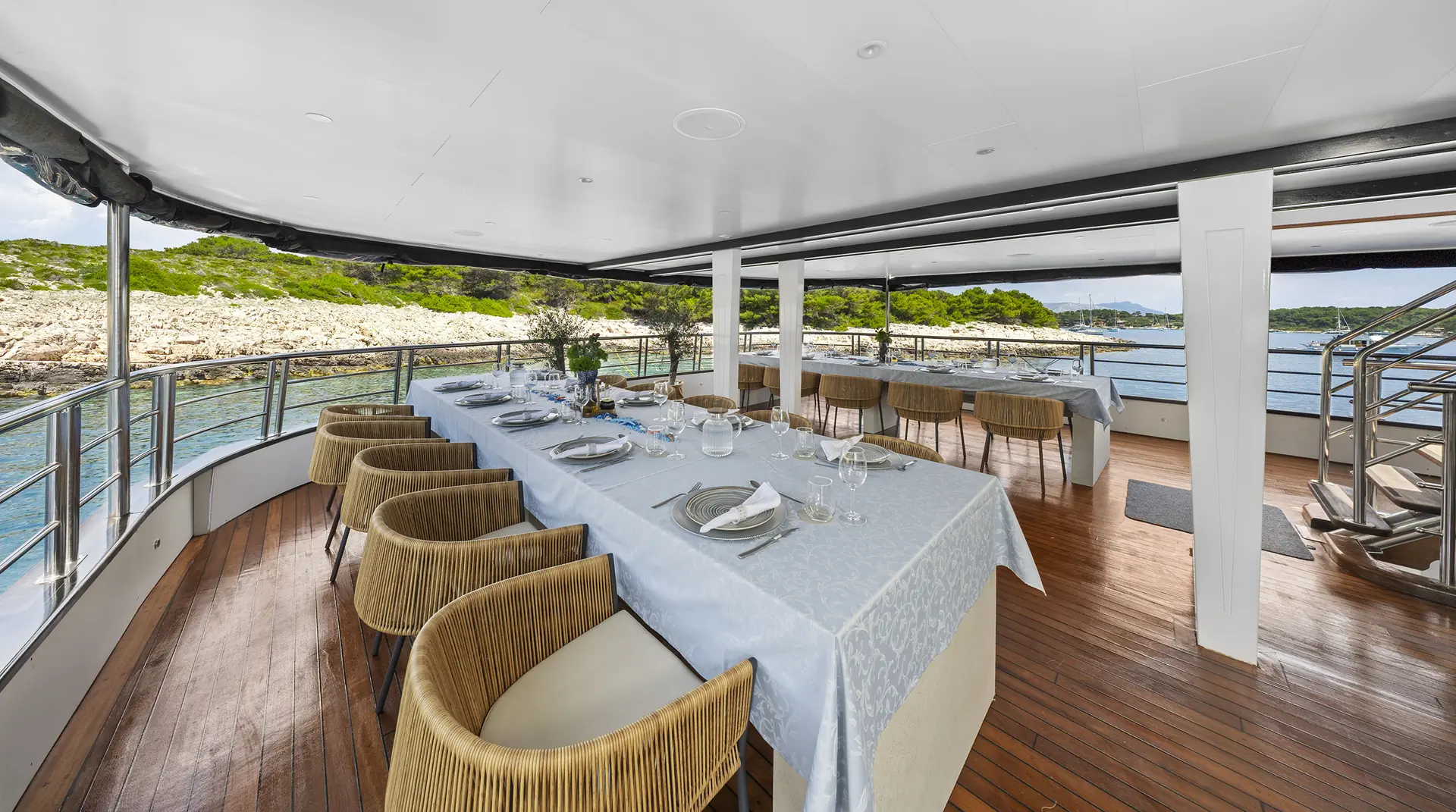 Elegant outdoor dining area on a yacht with a long table set for a meal, surrounded by beige chairs and overlooking scenic coastline with clear water and green trees, under a shaded deck with wood flooring.