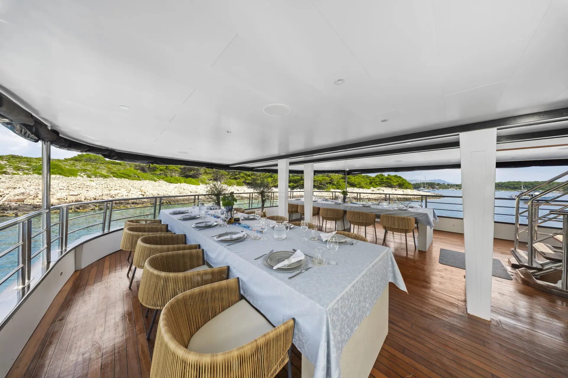 Outdoor dining area on a yacht with a long table set for a meal, surrounded by wicker chairs; ocean and rocky shoreline with greenery visible in the background.