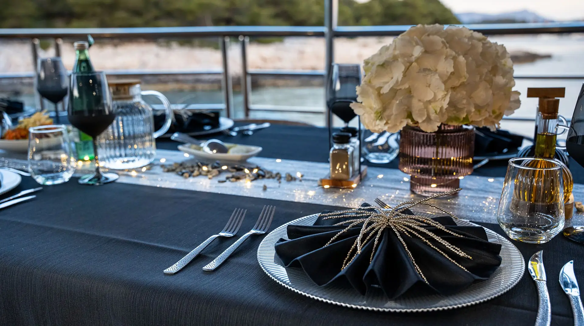 Elegant table setting on a boat with a black tablecloth, a decorative black napkin and silver charger, white flower centerpiece, wine glasses, and drinks, against a scenic water and shoreline background.