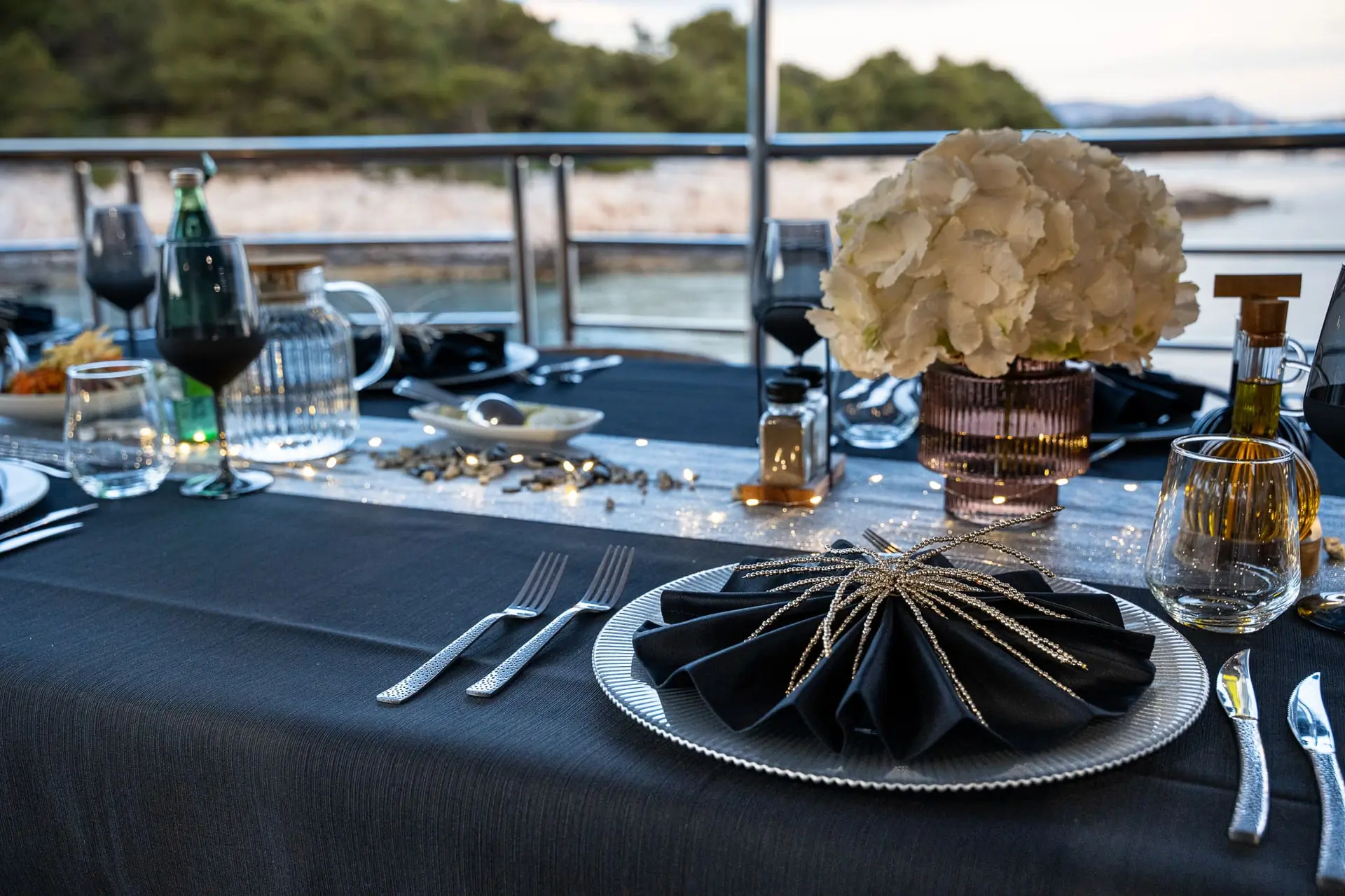 A close-up of an elegant table setting on a boat, featuring a black tablecloth, folded black napkin with gold decor, plates, glasses, cutlery, a vase of white flowers, and a scenic view of water and greenery in the background.